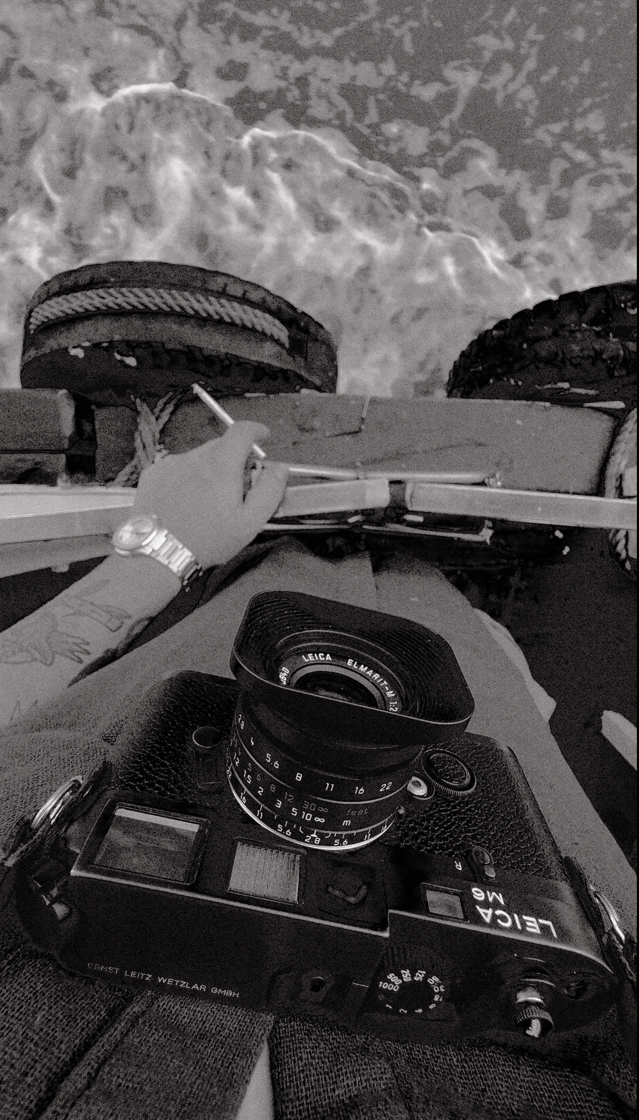 Fotografia in bianco e nero di una macchina fotografica Leica M6 appoggiata sulle gambe di una persona, con un obiettivo Leica Elmarit, e una mano con polsino di orologio che regge una tavola di legno con due ruote di bicicletta sopra un muro con il 