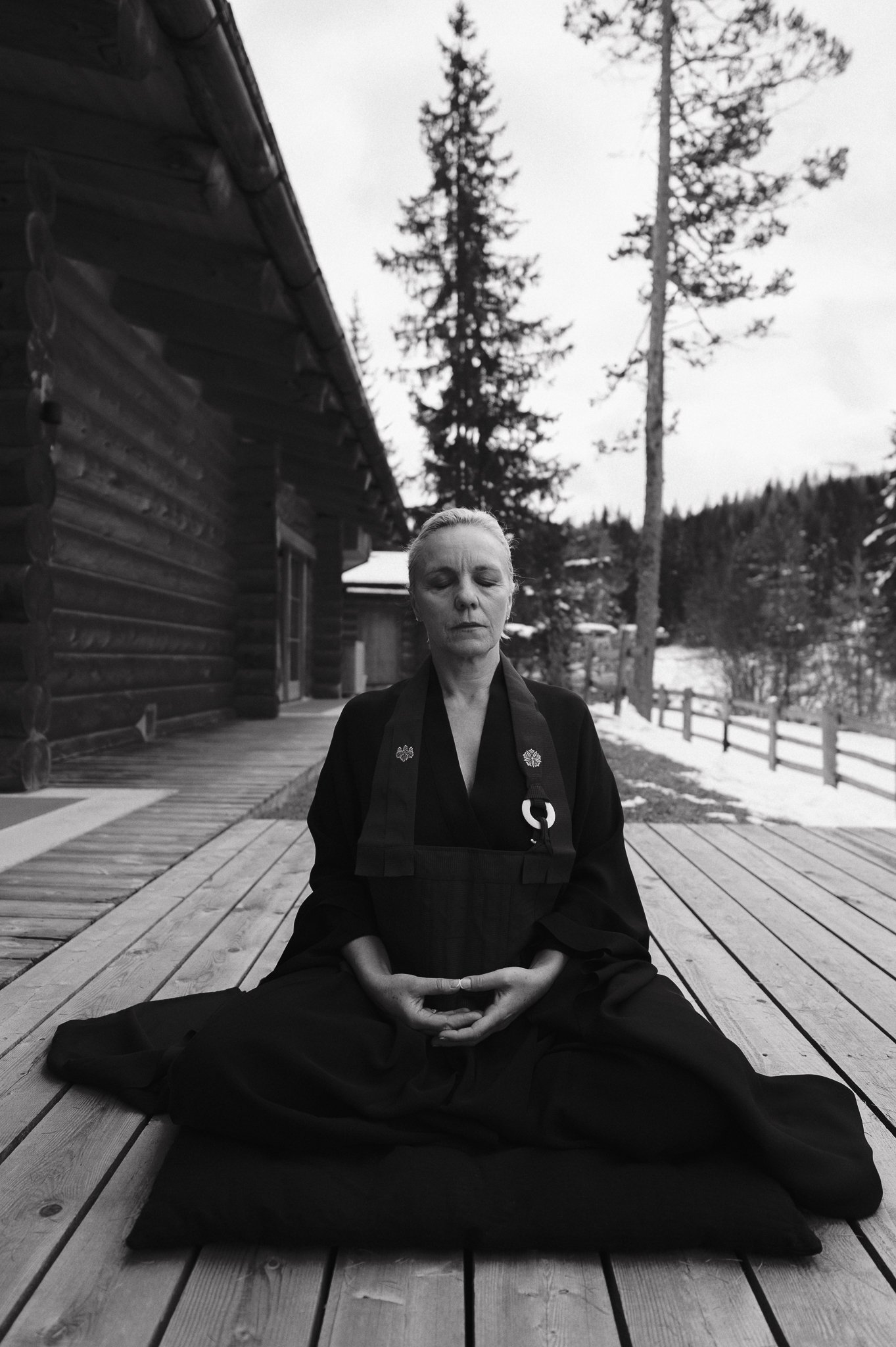 Una donna che pratica meditazione in posizione seduta con le gambe incrociate su un tappetino nero sulla terrazza di legno davanti a un edificio in legno, con alberi e neve sullo sfondo.