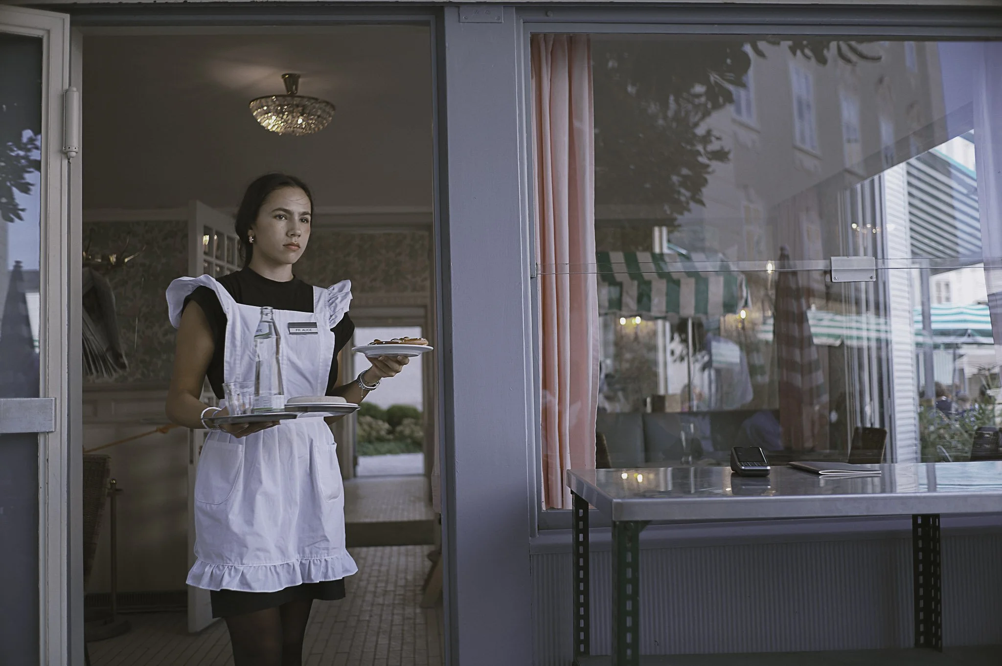 Una cameriera con grembiule bianco e vestito nero serve cibo in un ristorante o caffetteria, vista attraverso una porta di vetro