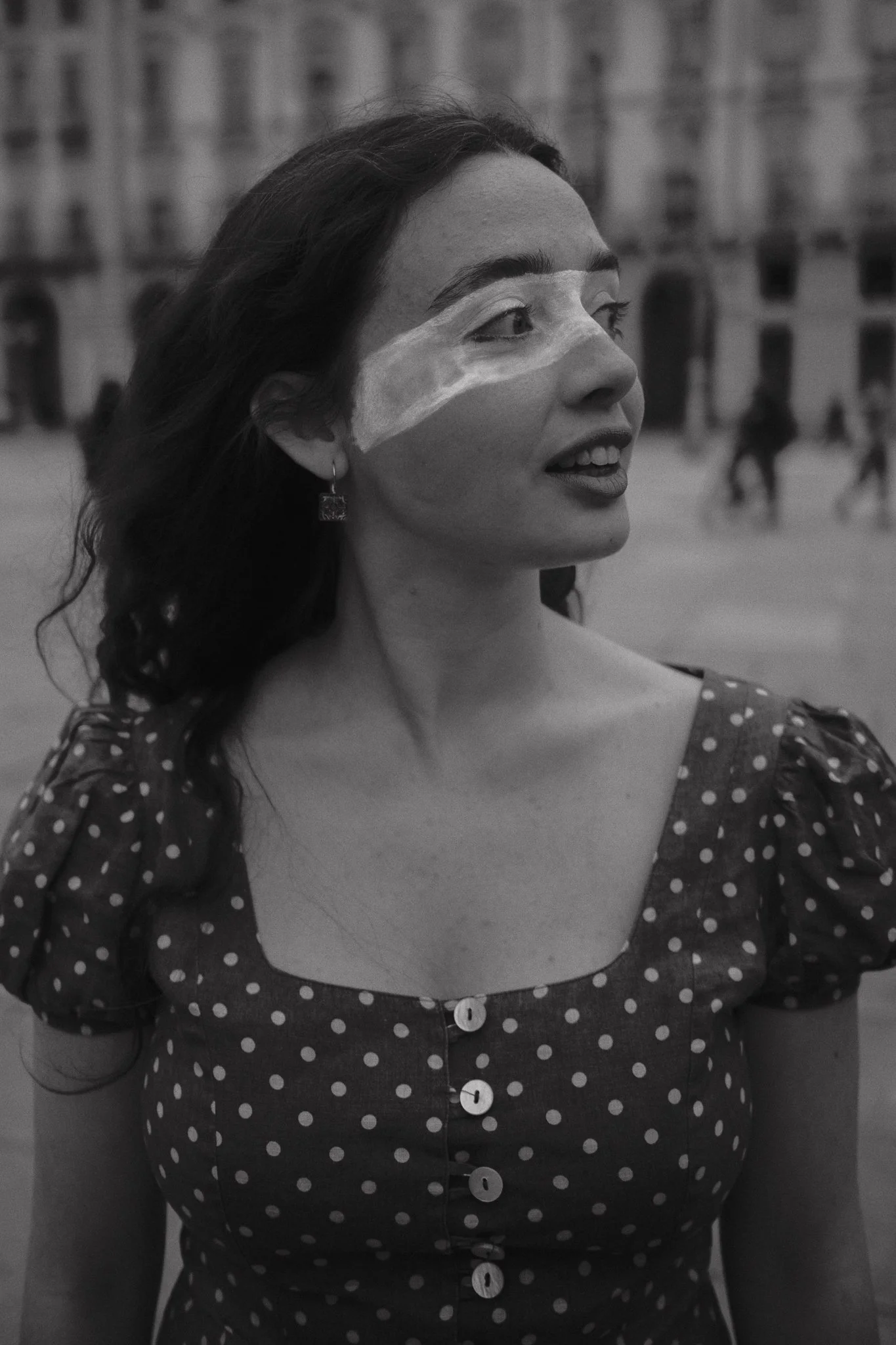 Una donna con capelli lunghi e ricci, indossa un vestito a maniche corte con motivo a pois e una tape sulla faccia, si trova in una piazza con edifici sullo sfondo.