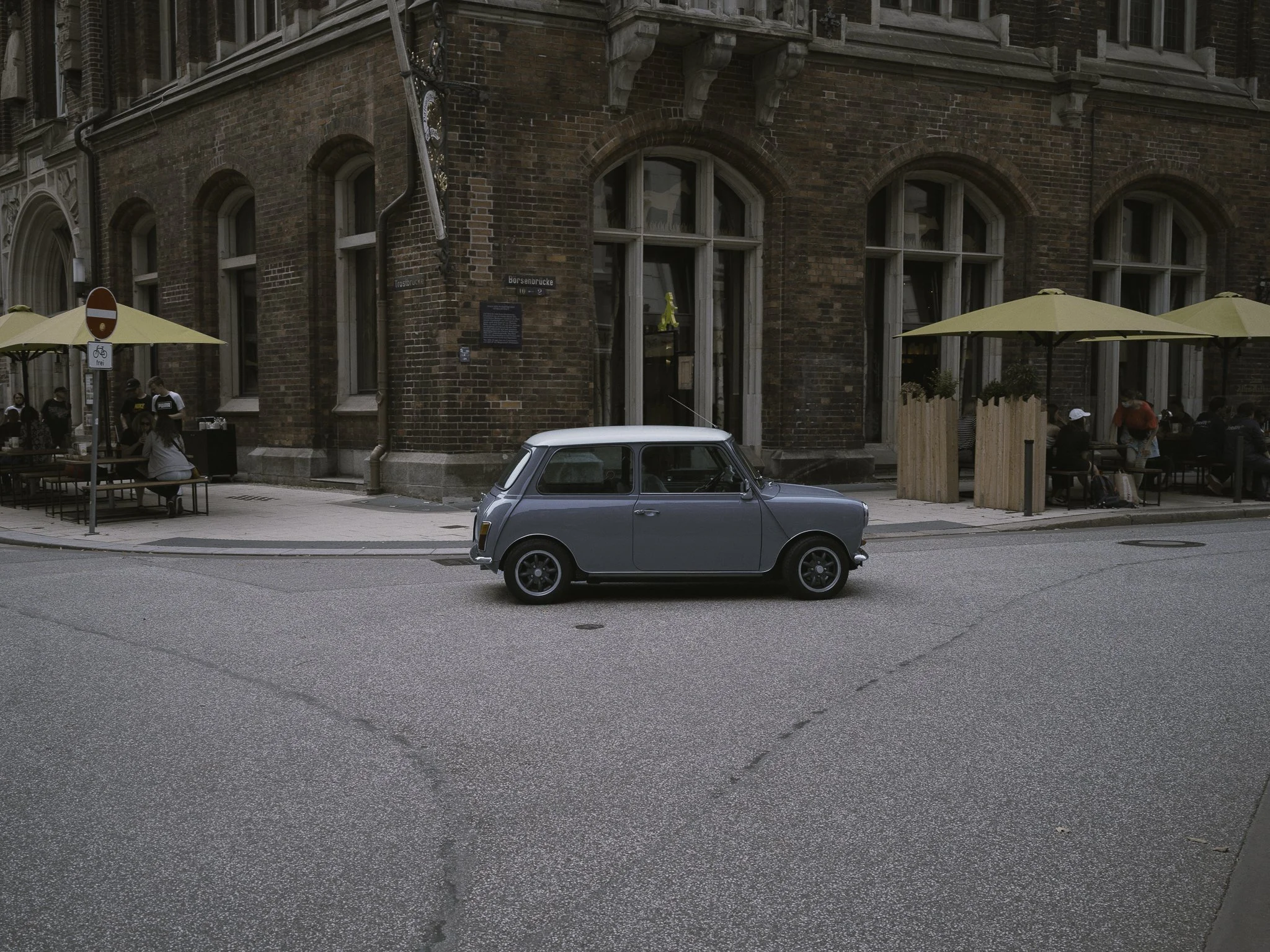 Una strada cittadina con un'auto compatta di color grigio parcheggiata, sullo sfondo un edificio in mattoni con finestre grandi e un'area di ristorazione all'aperto con sedie, tavoli e ombrelloni gialli.