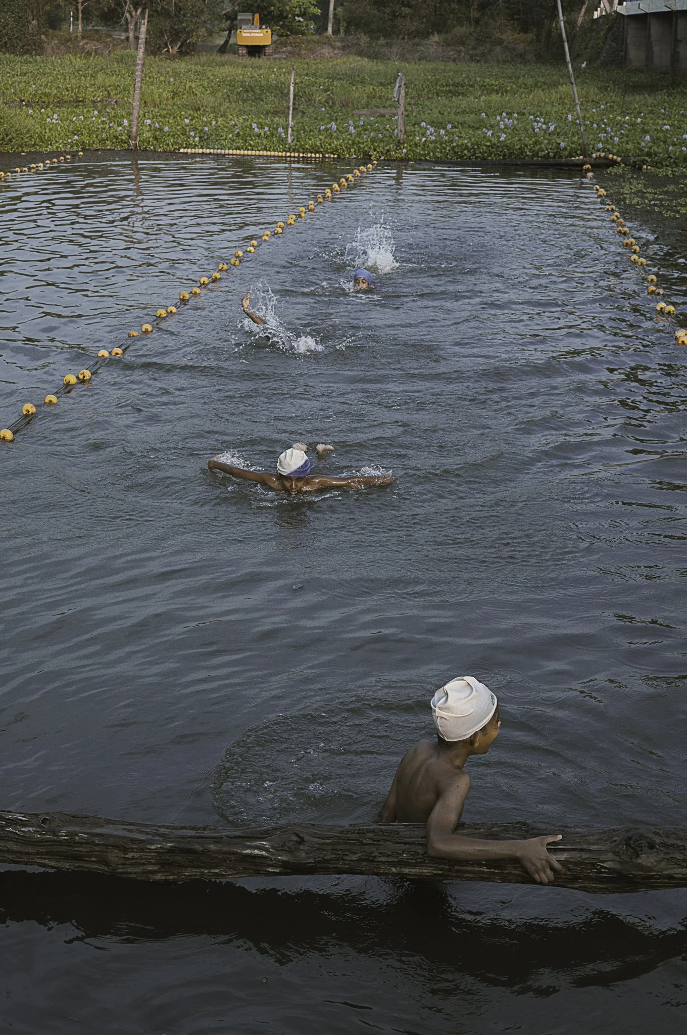 Bambini che nuotano in una piscina all'aperto delimitata da boe arancioni, con uno di loro che si appoggia a un tronco sul bordo della piscina.