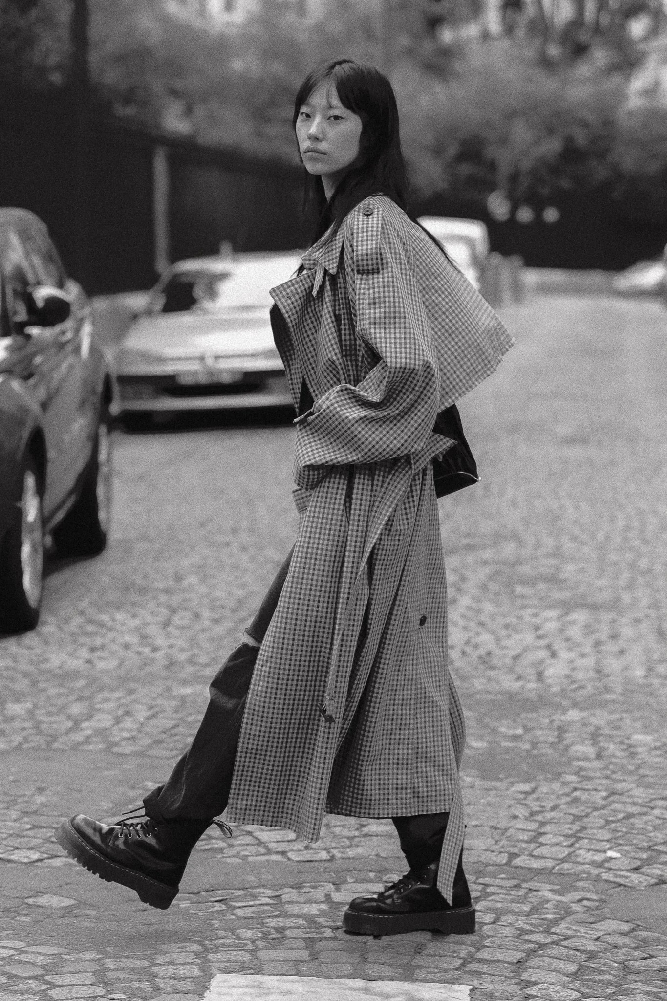 Una donna vestita con un completo check in stile streetwear, con scarpe nere robuste, cammina sulla strada in un'area urbana con auto parcheggiate sullo sfondo, in bianco e nero.