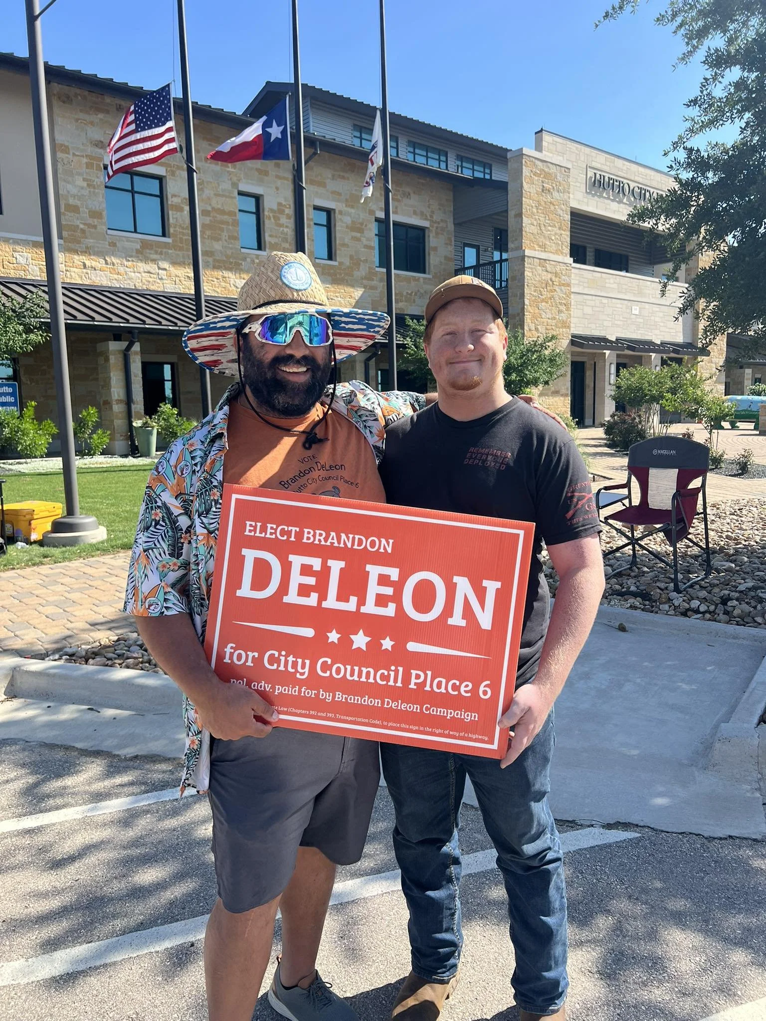 Two men standing outdoors holding a campaign sign for Brandon DeLeon running for City Council Place 6. One man is wearing a broad-brimmed hat, sunglasses, and a colorful shirt, and the other is wearing a black T-shirt and a cap. There is a building with flags and a sign that says 'Hutto City' in the background.