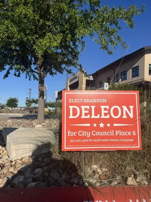 A red campaign sign for Brandon Deleon, running for City Council Place 6, is displayed outdoors near a tree, with a building and clear blue sky in the background.
