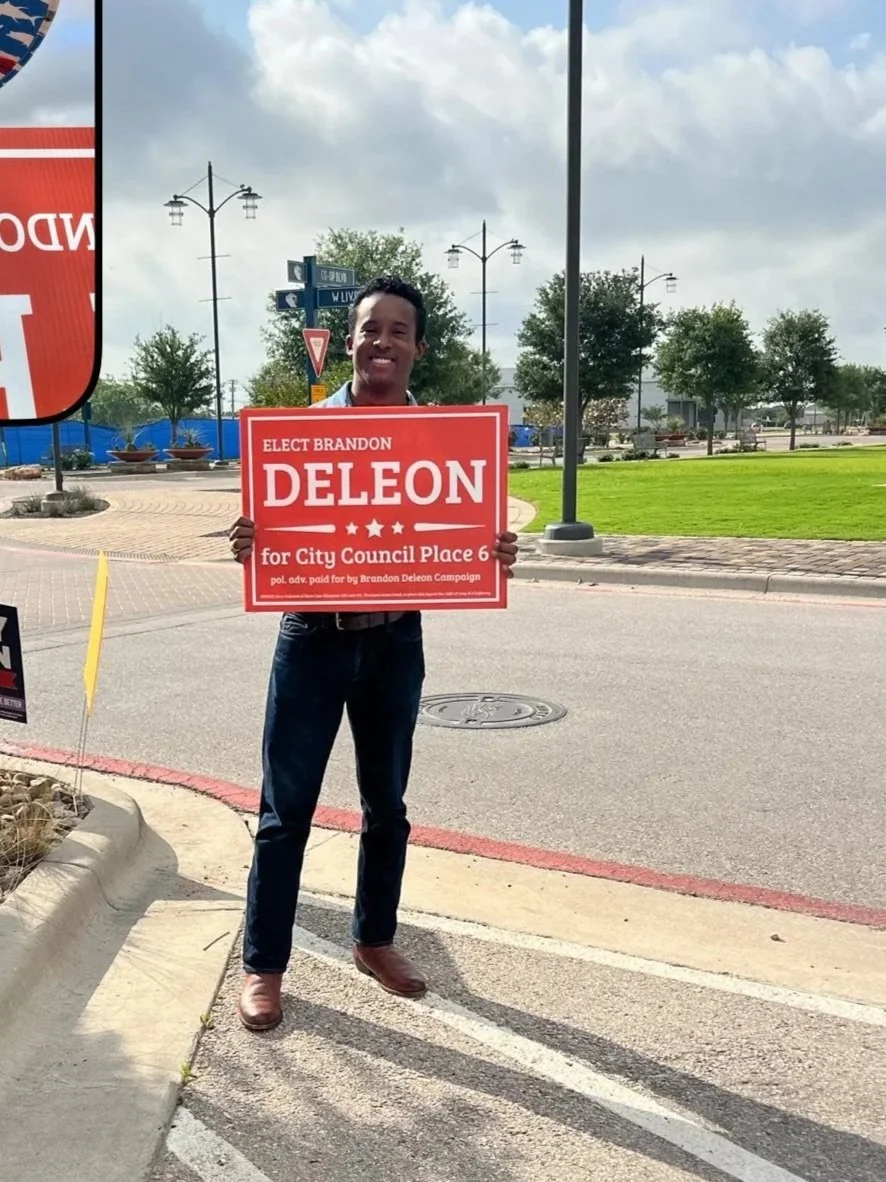 A man standing outdoors on a sidewalk holding a political campaign sign that reads 'Elect Brandon Delon for City Council Place 6'. The background includes trees, street lamps, traffic signs, and a partly cloudy sky.