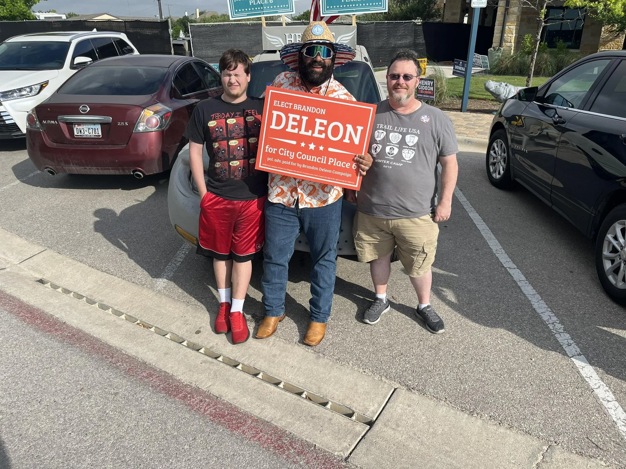 Three men standing in a parking lot, holding a campaign sign that reads 'Elect Brandon Deleon for City Council Place 6.' The man in the middle is wearing a large sombrero and sunglasses, and the man on the right is wearing a gray T-shirt with various logos. The man on the left is wearing a black T-shirt with red shorts. Several cars are parked behind them.