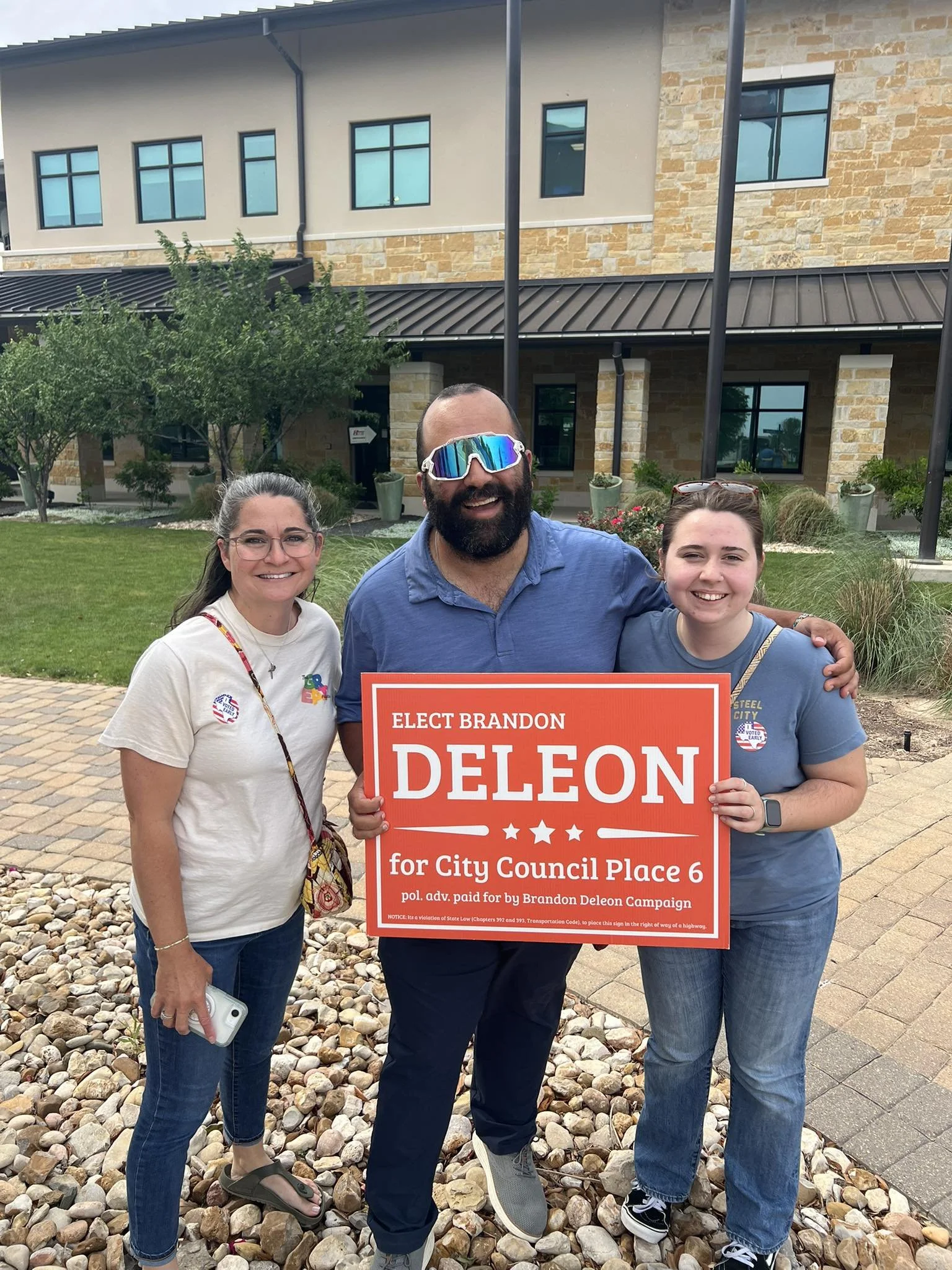 Three people standing outside in front of a building, holding a red campaign sign that reads "Elect Brandon Deleon for City Council Place 6." The man in the middle is wearing sunglasses and has a beard, while the two women on either side are smiling and wearing casual clothes.