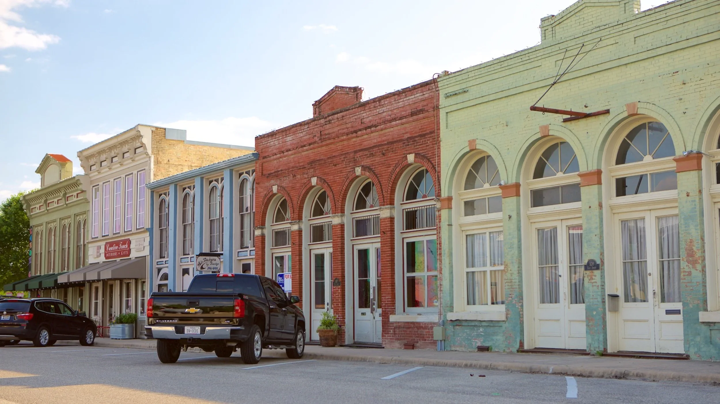 A row of small historic storefronts painted in pastel colors, with parked cars in front on a sunny street.