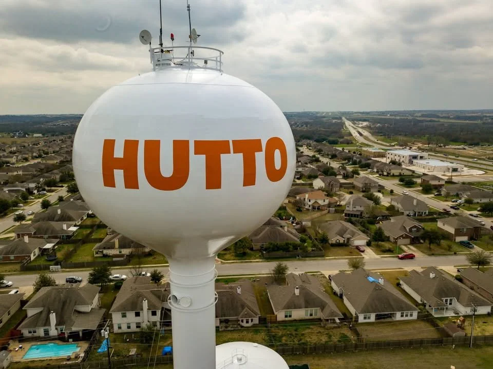 Large white water tower with orange letters spelling 'HUTTO' in a residential neighborhood with houses, streets, and a cloudy sky visible in the background.