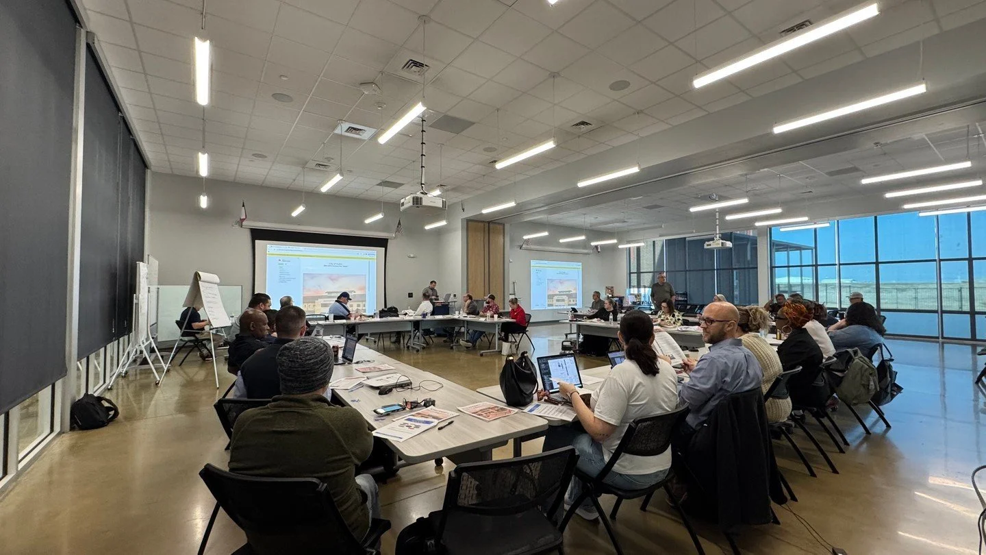 A diverse group of people attending a meeting or workshop in a modern conference room with large windows, presentation screens, and seating arranged in a U-shape.