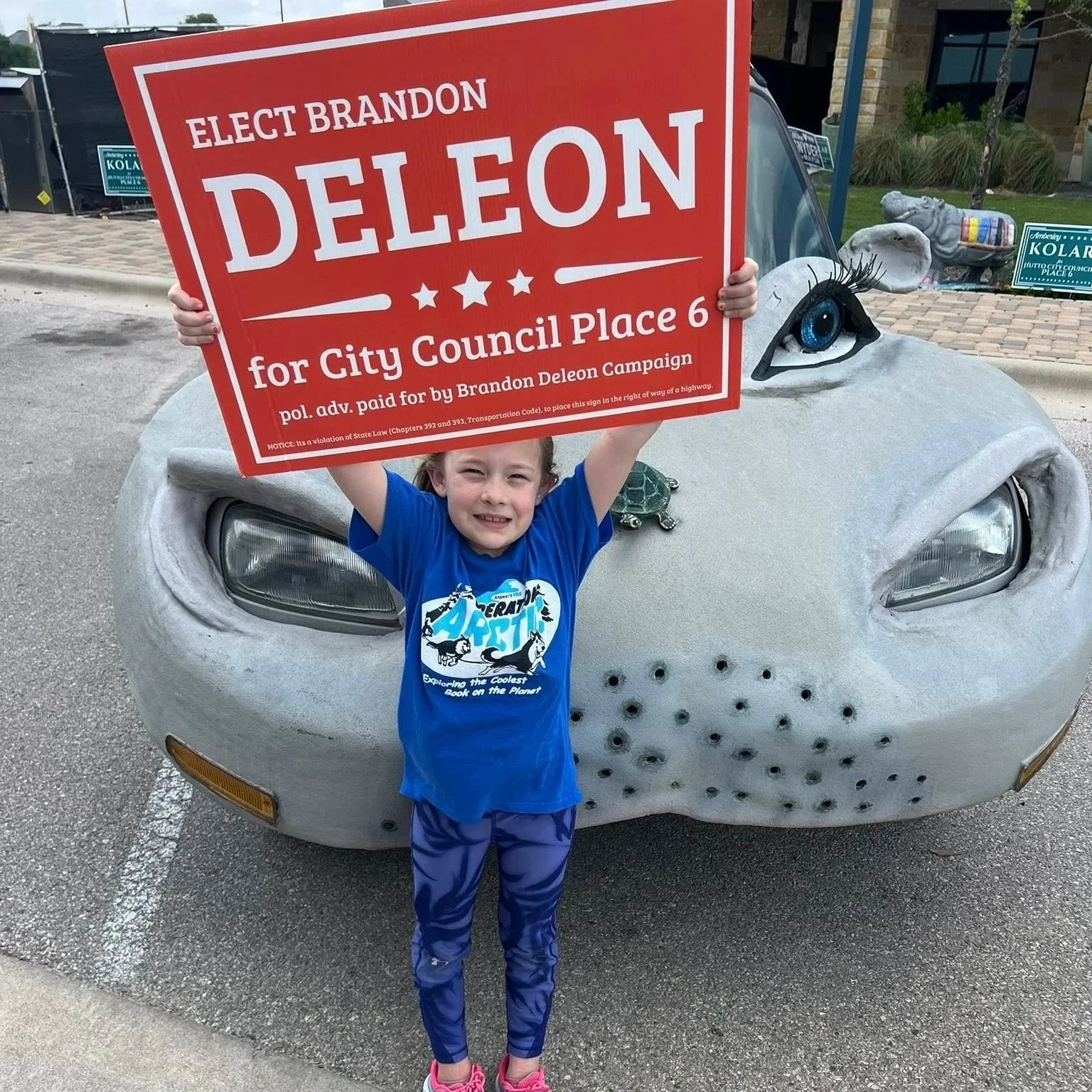 A young girl holding a red campaign sign in front of a car with a painted whale face. The sign reads "Elect Brandon Deleon for City Council Place 6." She is smiling and wearing a blue shirt with a cartoon dog on it and blue leggings.