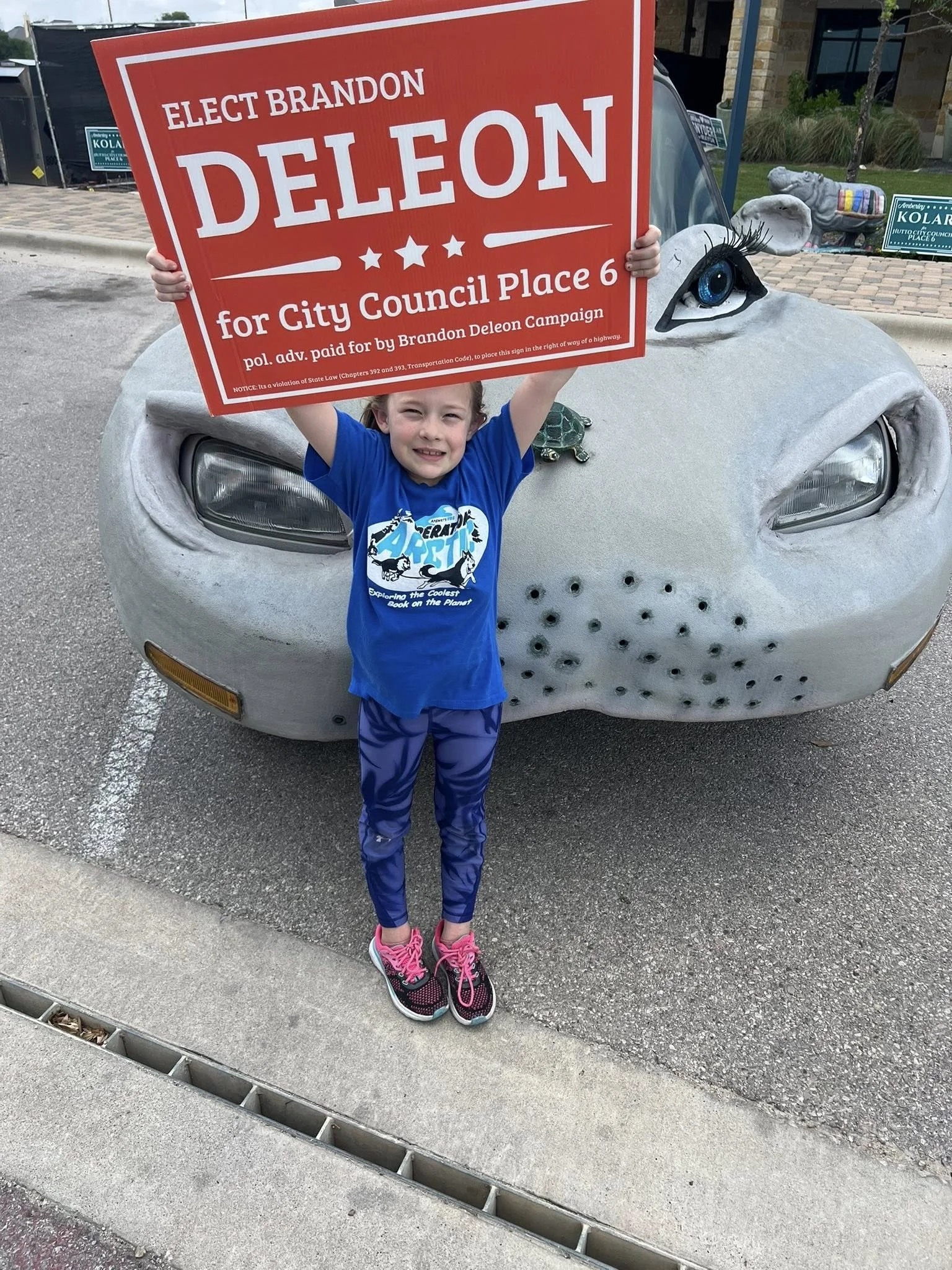 A young girl with a big smile holding a campaign sign for Brandon Deleon in front of a car painted to look like a turtle. The sign reads 'Elect Brandon Deleon for City Council Place 6' and mentions campaign funding.