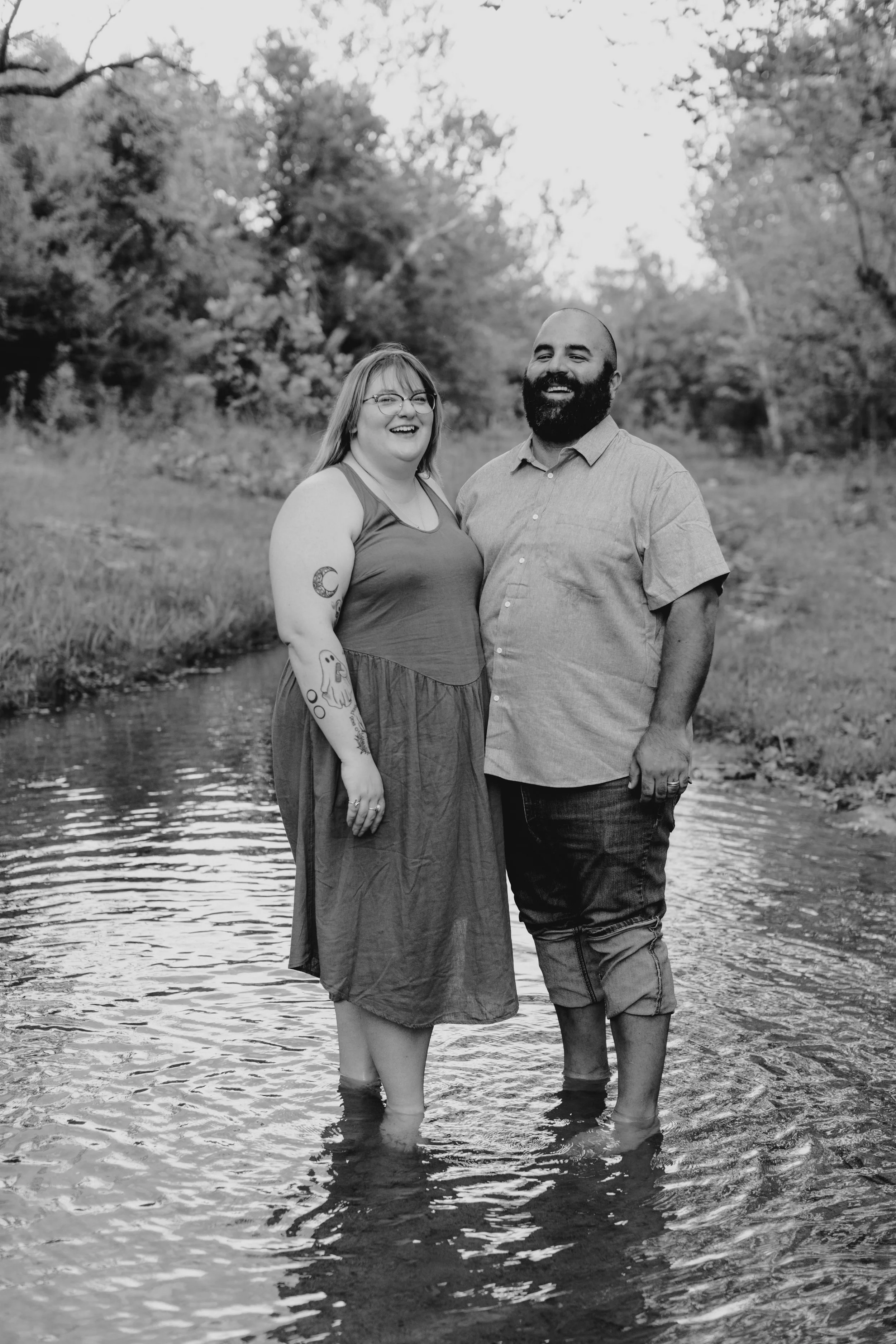 A smiling couple standing in a creek with water up to their ankles, surrounded by trees and nature, in a black and white photograph.