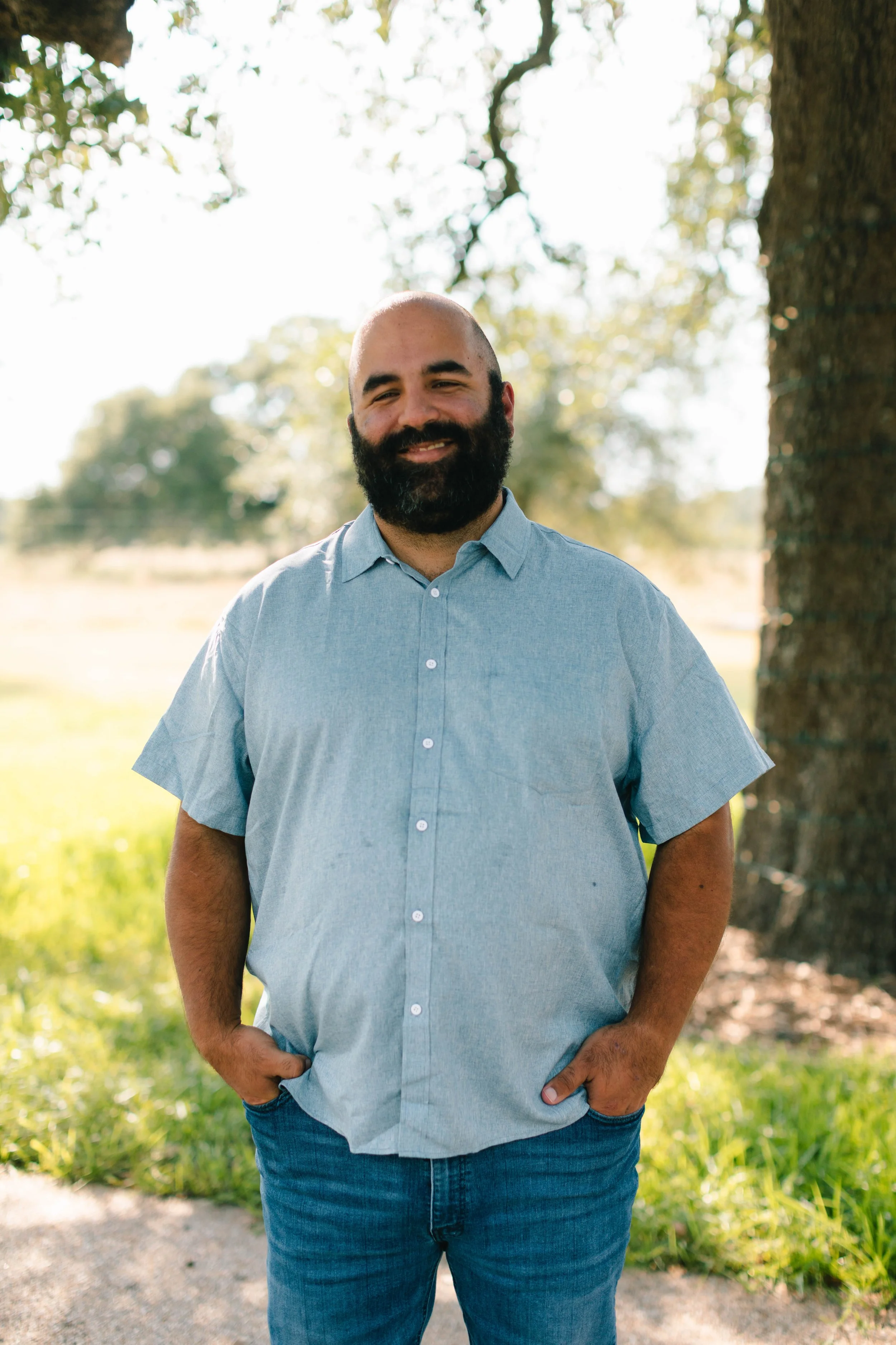 A man with a beard standing outdoors near a tree, wearing a light blue short-sleeved shirt and jeans, smiling at the camera with hands in pockets.