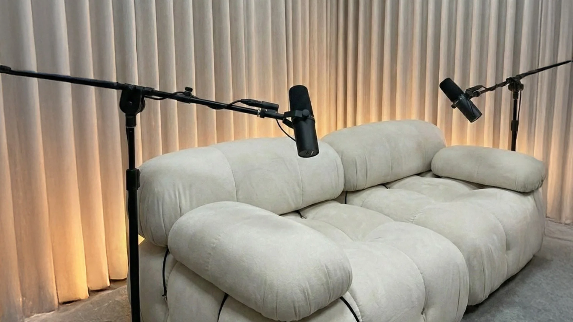 A cream-colored sectional sofa with plush cushions in front of beige curtains, with two black microphones on stands positioned near the sofa.