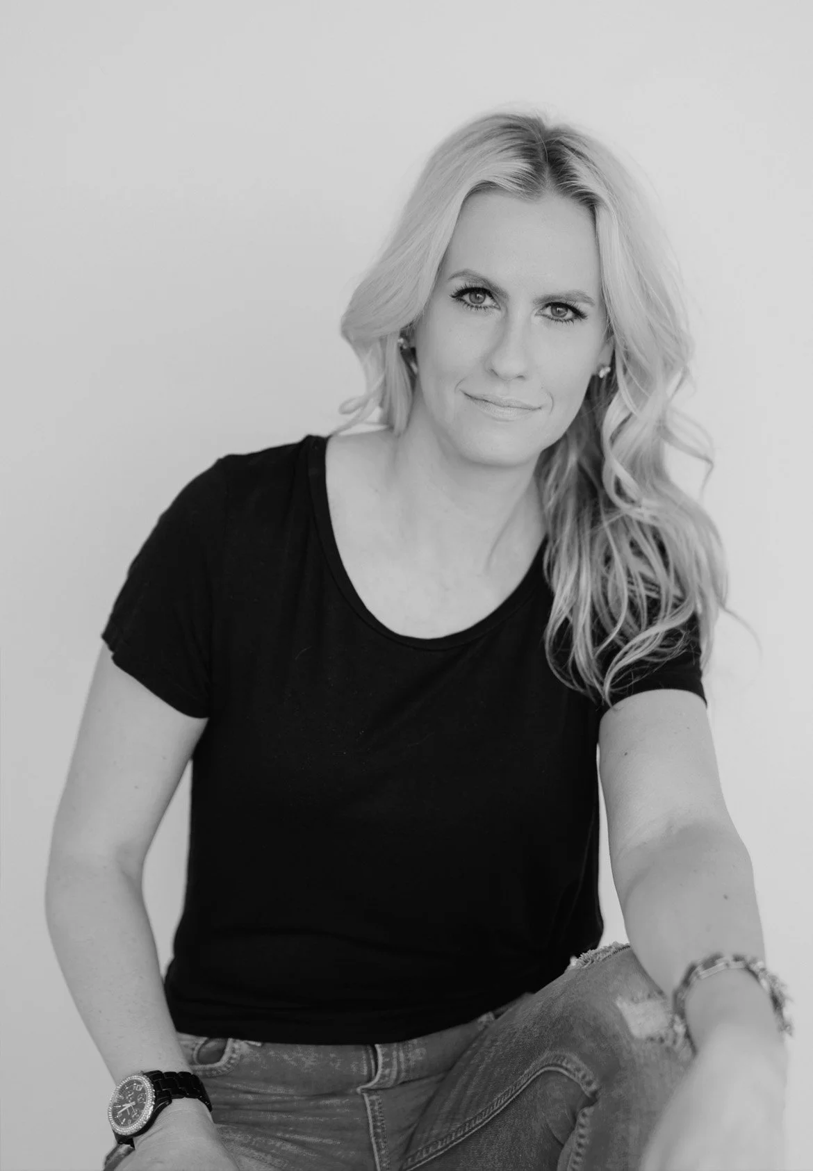 Black and white portrait photo of a woman with wavy blonde hair, wearing a black t-shirt, sitting against a plain light background.