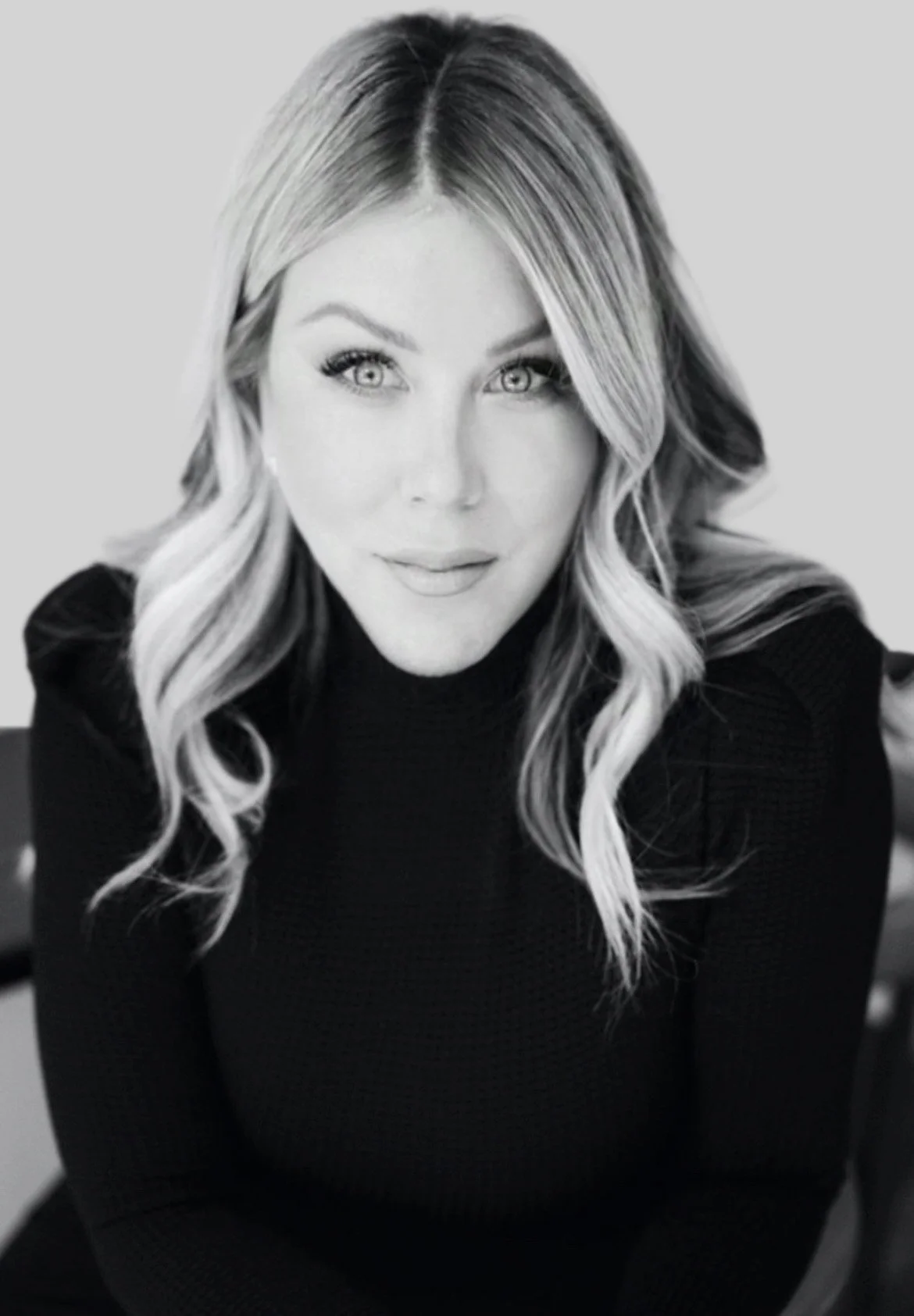A black-and-white portrait of a woman with wavy, shoulder-length hair, wearing a dark textured turtleneck sweater, looking directly at the camera with a slight smile.