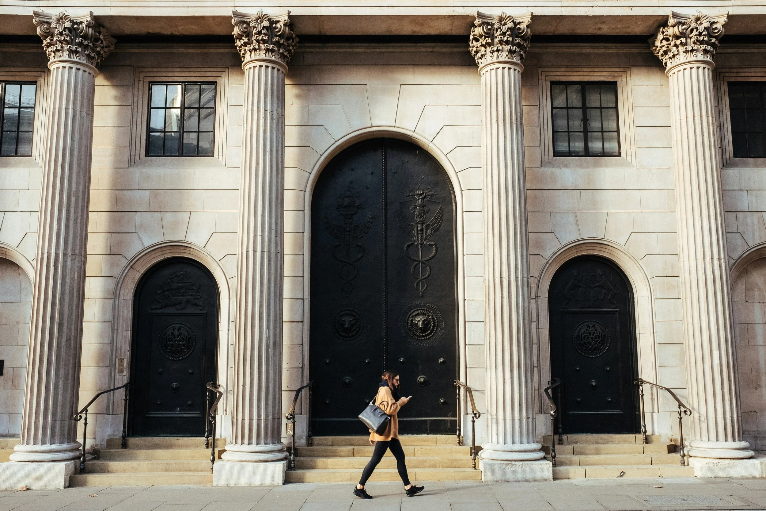 A woman walking past the front of a neoclassical building with large decorative columns and three black doors, with intricate carvings including symbols of medicine and law.