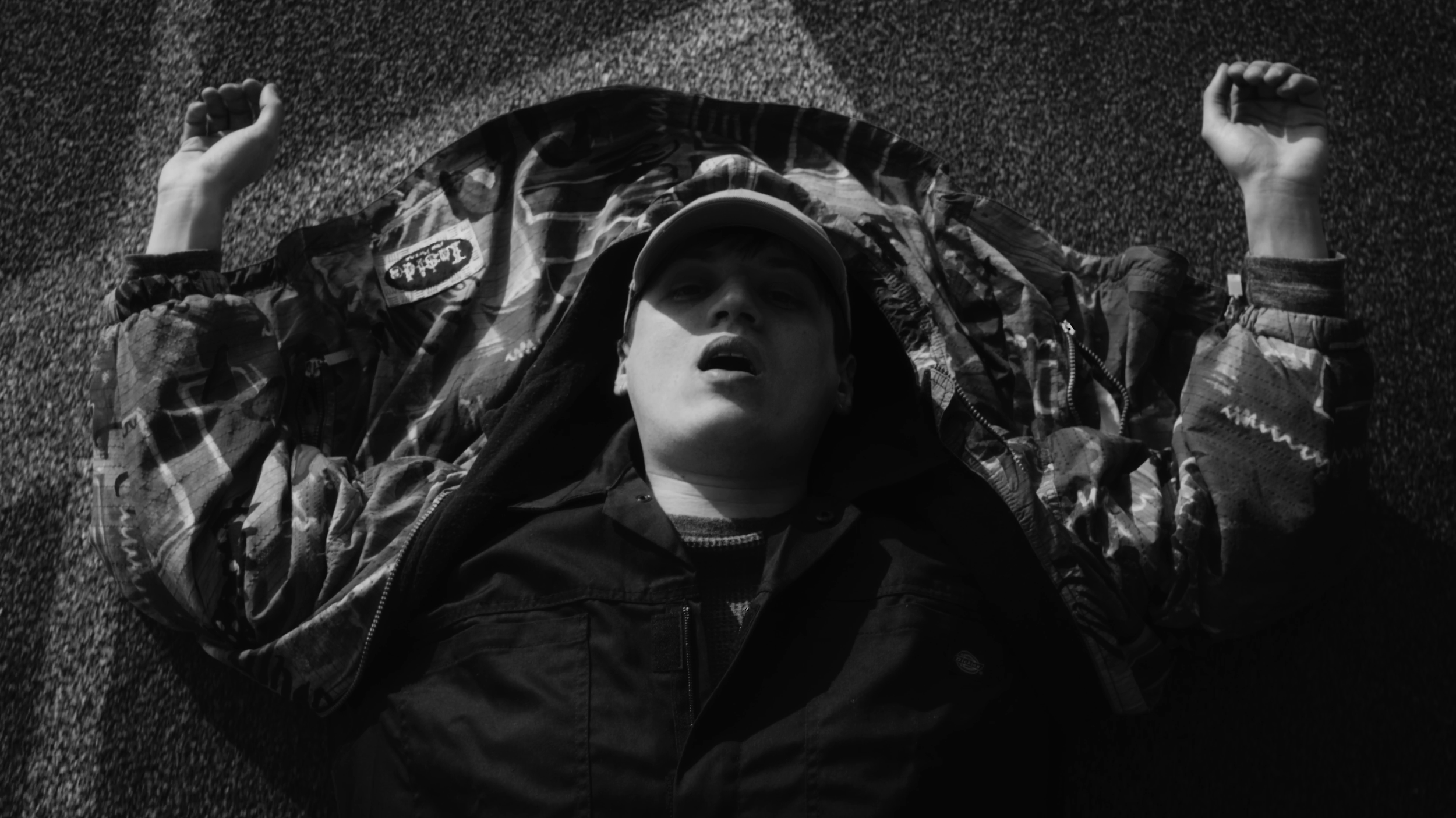 Jeune homme allongé sur le sol, vu de dessus, avec bras tendus, portant une casquette et une veste déstructurée, dans une ambiance sombre.