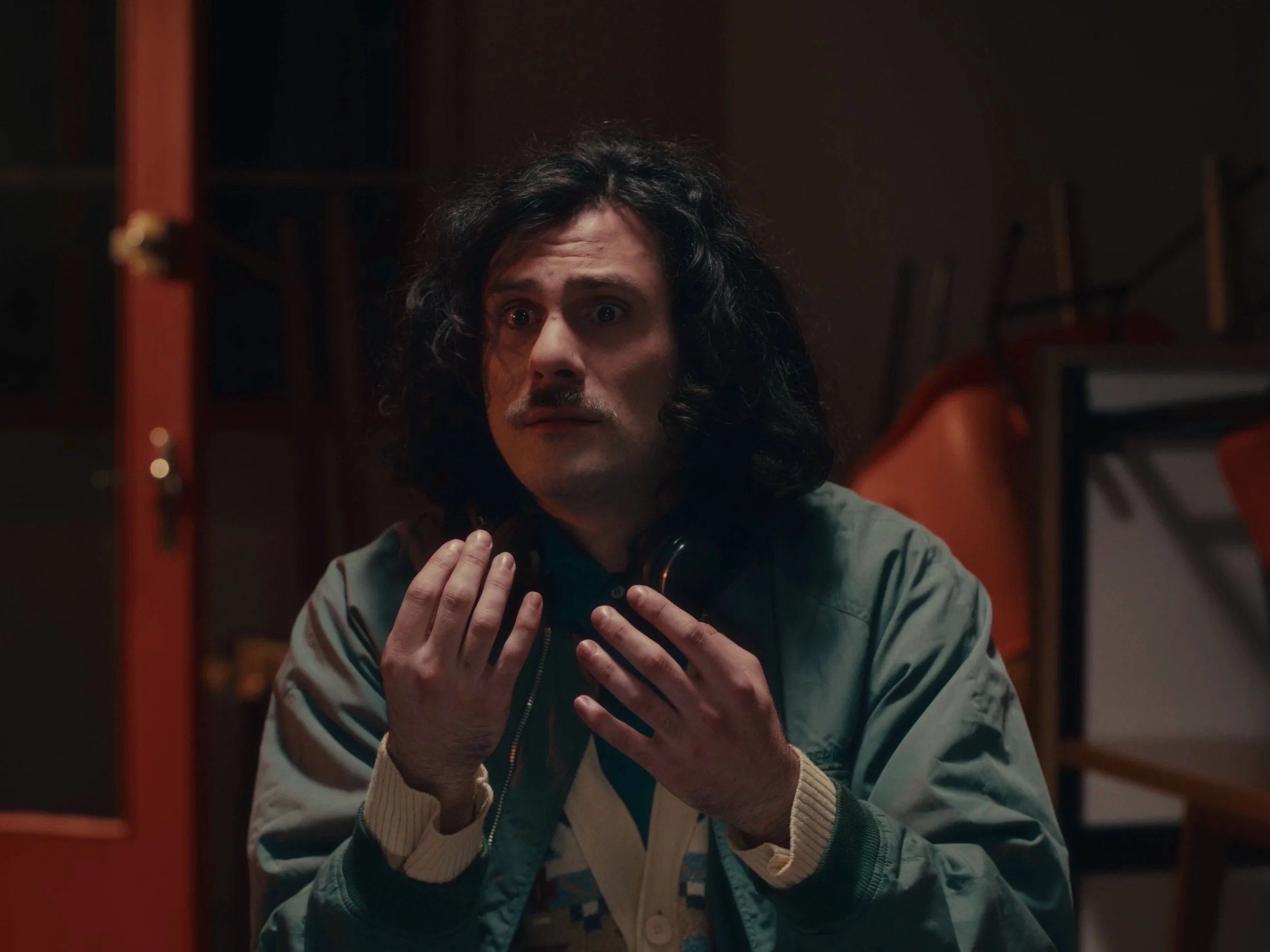 Un homme avec de longs cheveux noirs, portant un casque autour du cou, semble surpris ou inquiet, dans un espace intérieur avec des meubles oranges en arrière-plan.