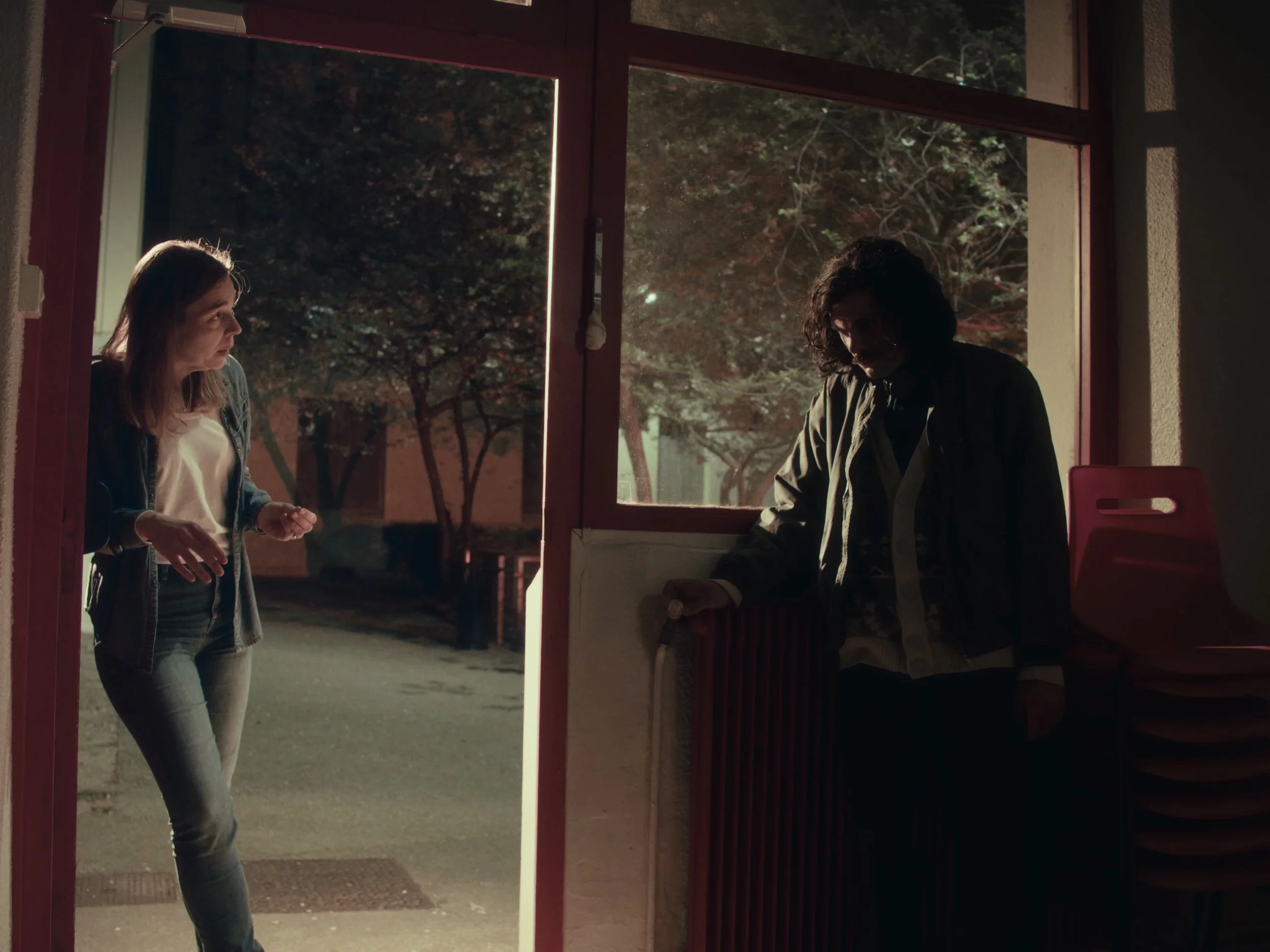 Une femme discutant avec un homme à l'intérieur d'une pièce proche d'une porte donnant sur l'extérieur, avec des arbres visibles. La scène est sombre avec une lumière naturelle venant de l'extérieur.