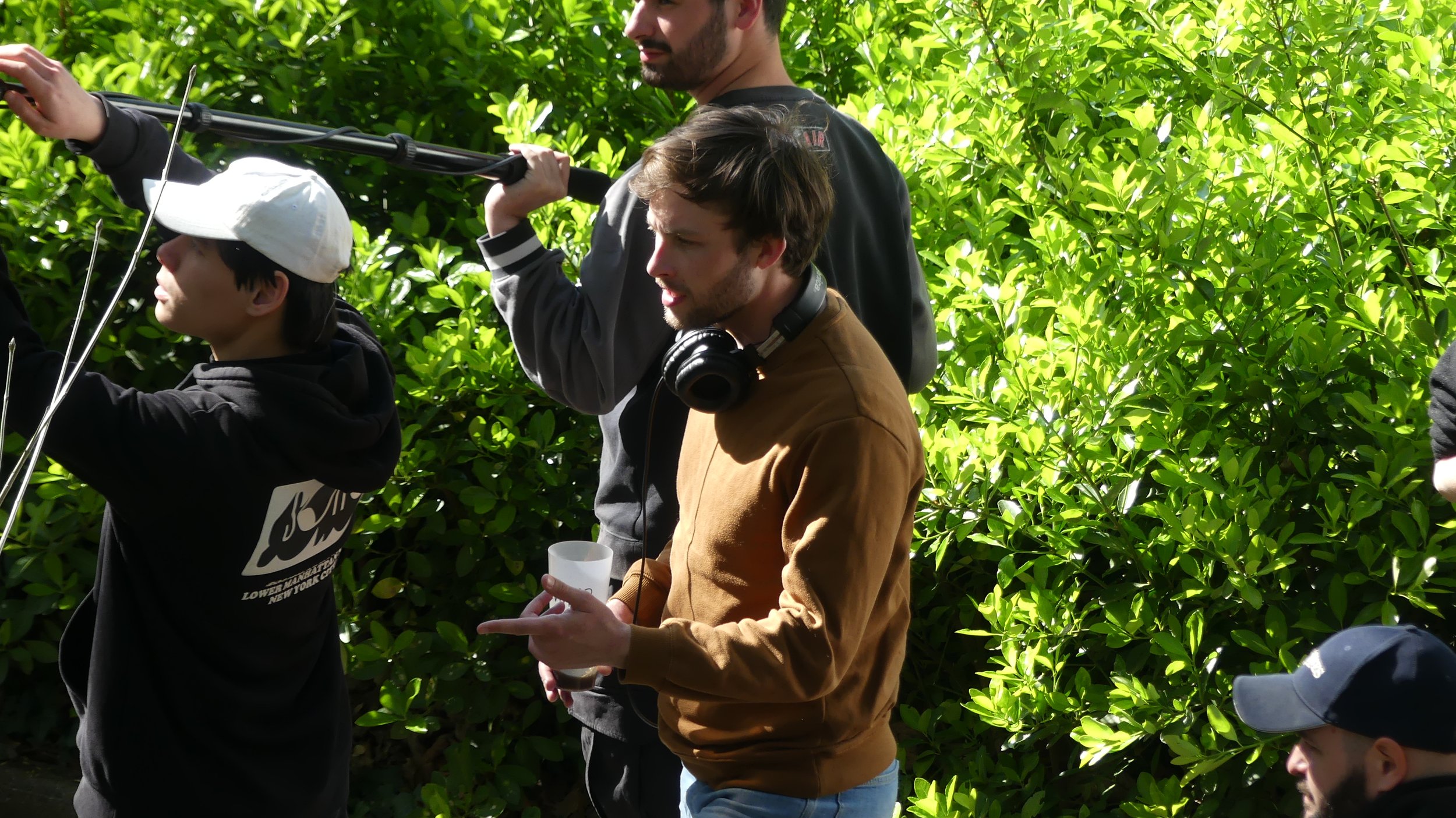 Groupe de personnes en plein air devant des arbustes verts, dont un homme porte un casque audio et tient un gant en plastique, un autre homme porte une veste grise avec un casque autour du cou, une personne avec un hoodie noir et une casquette blanche, et une autre personne avec une casquette noire.