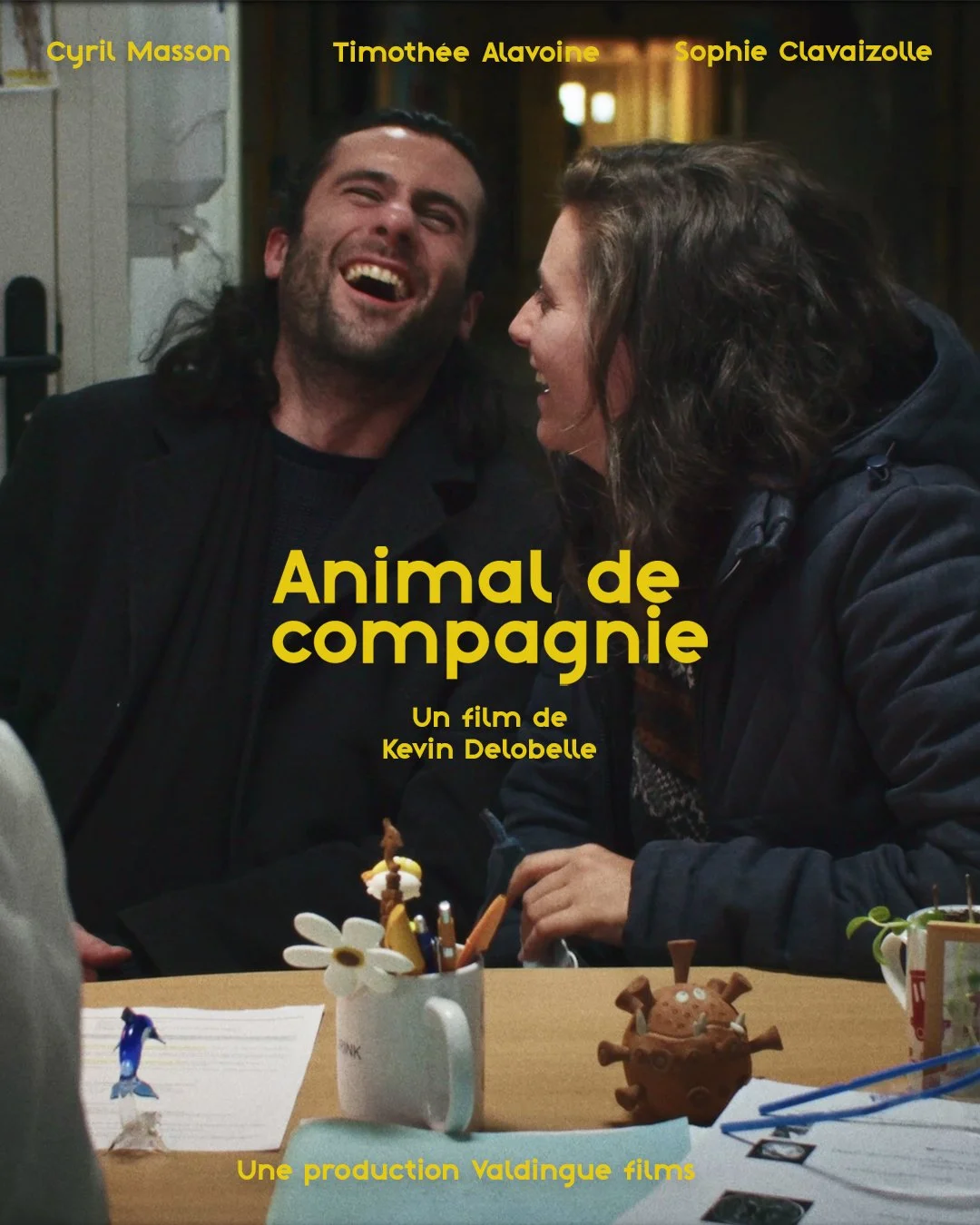 Une affiche de film intitulée "Animal de compagnie" avec deux personnes qui rient et discutent, assises à une table avec des objets, un mug avec des fleurs en plastique, et un modèle de virus en chocolat.
