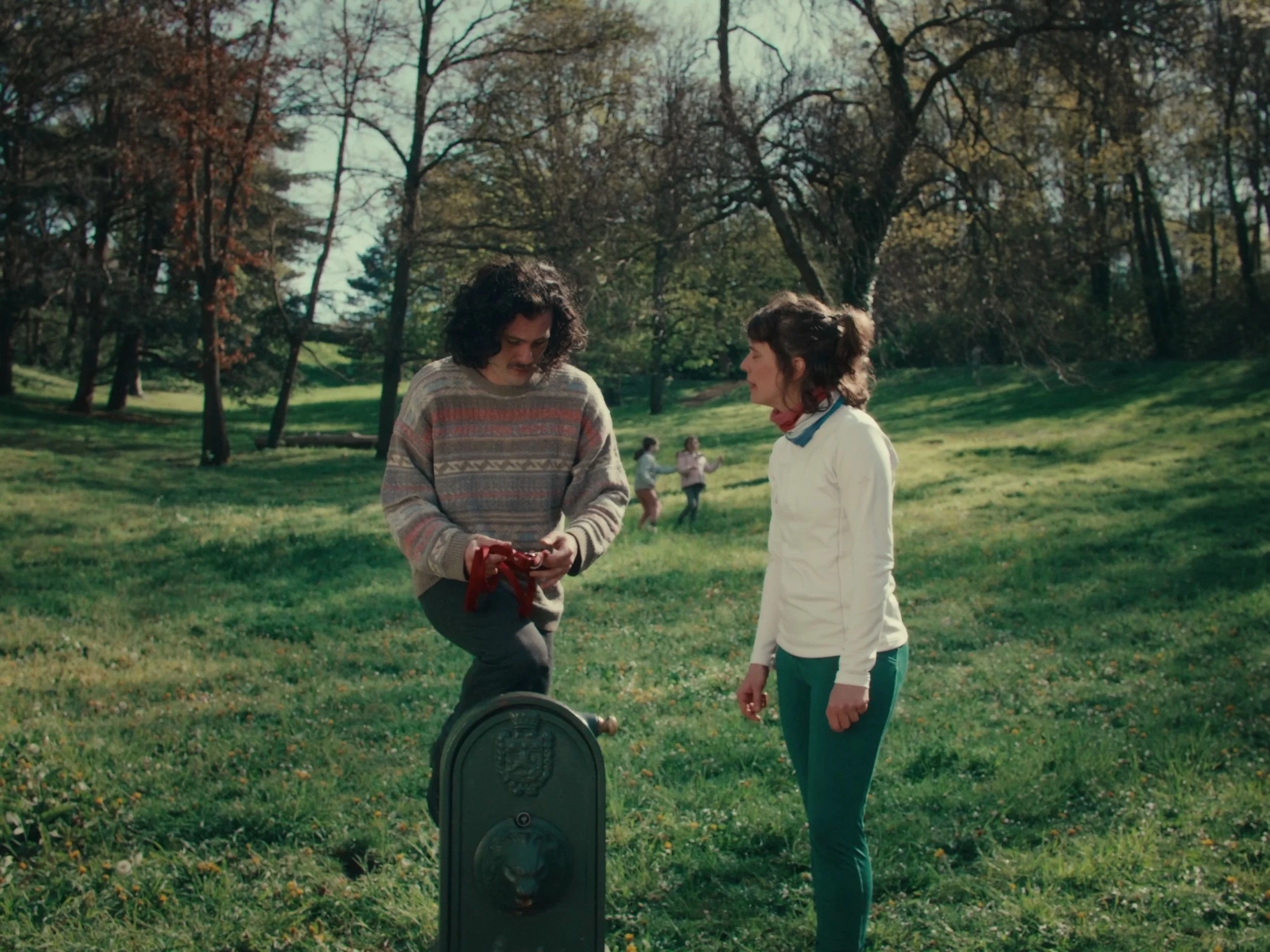 Une scène dans un parc avec deux femmes en conversation, un homme à gauche tenant un objet rouge et une boîte aux lettres en avant, d'autres personnes en arrière-plan.