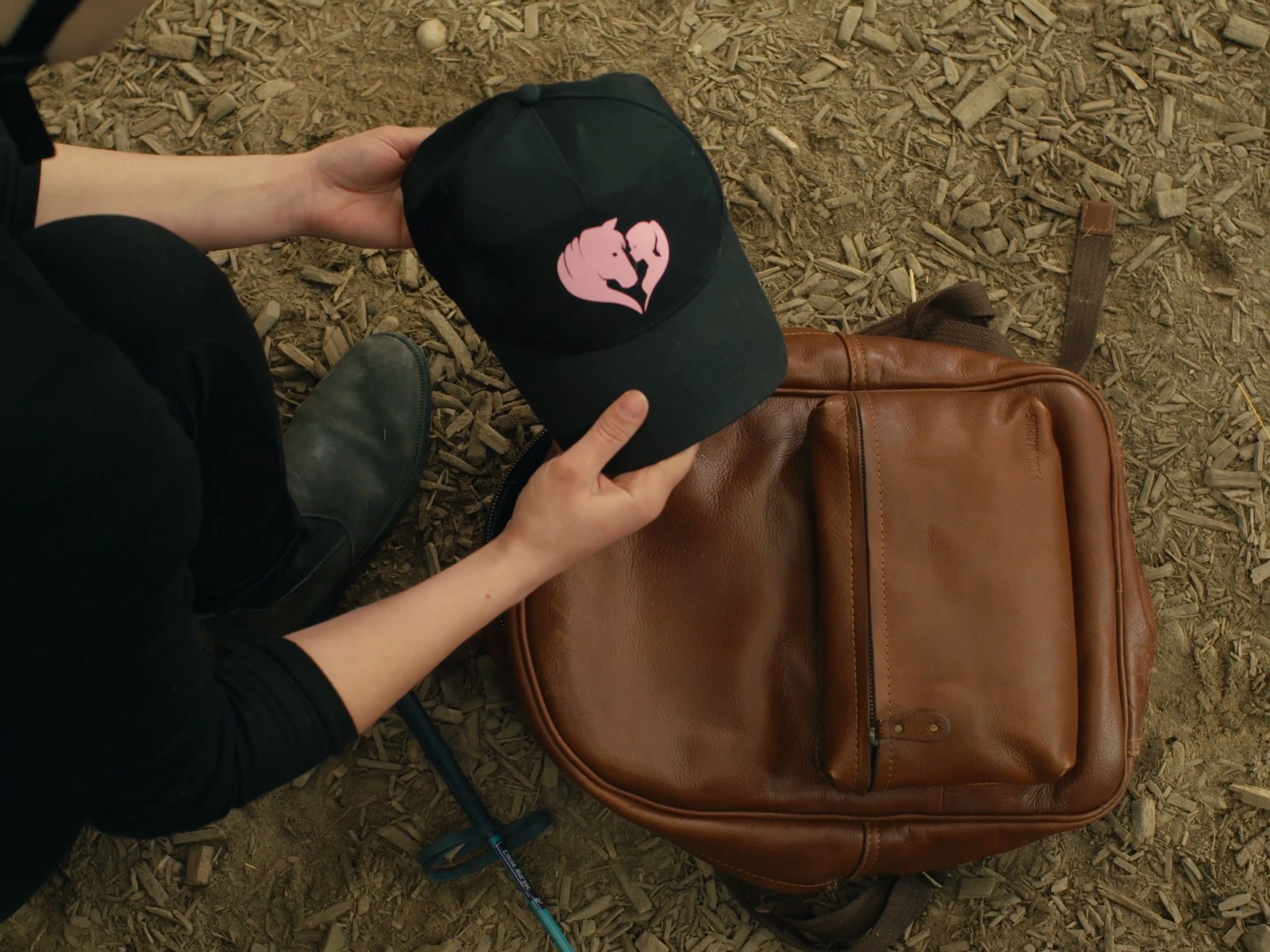 Une personne tient une casquette noire avec un logo de deux flamants roses formant un cœur, posée sur un sac en cuir marron, sur une surface de sol en bois.