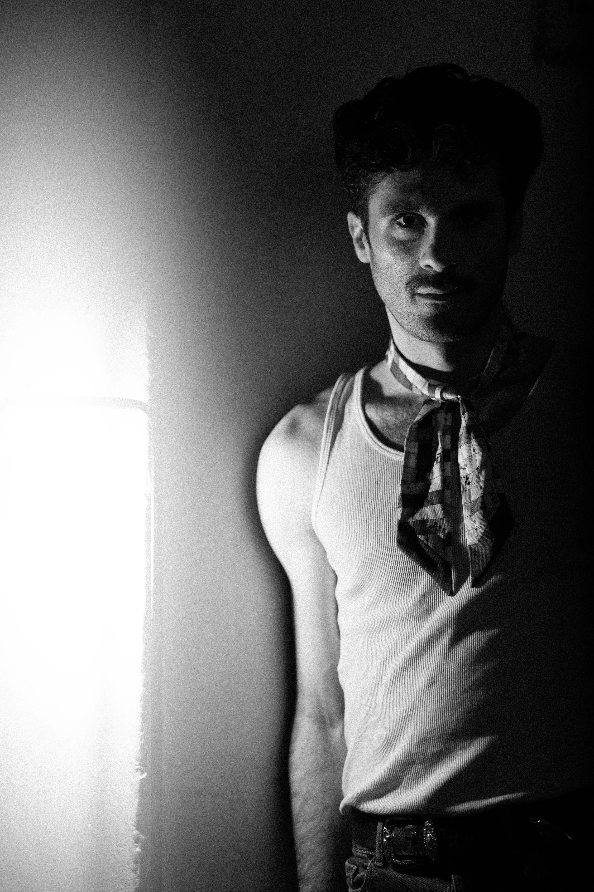 Portrait en noir et blanc d'un homme avec un bandana autour du cou, portant un débardeur, debout devant un mur, moitié gauche éclairee, moitié droite dans l'ombre.