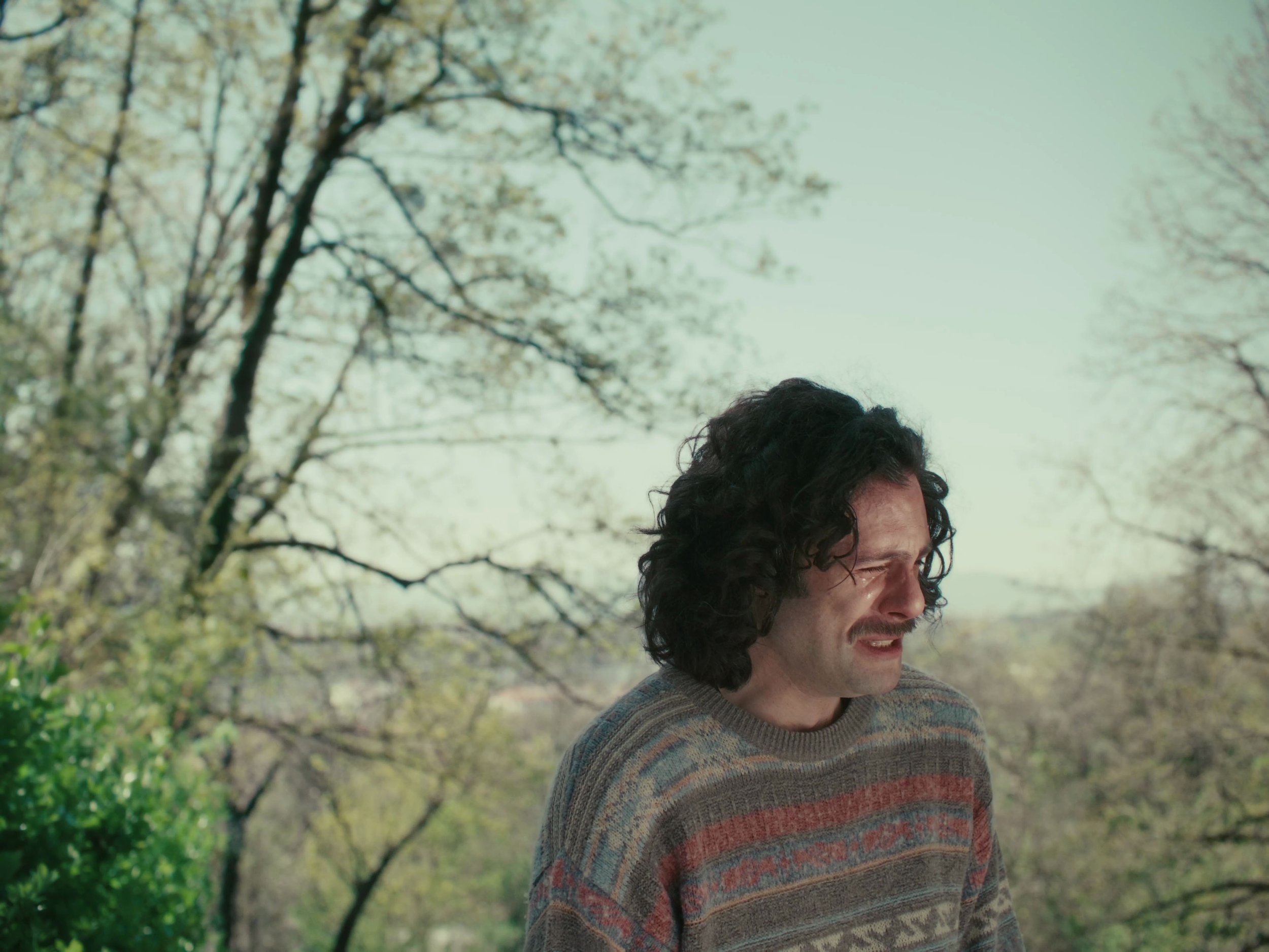 Homme avec des cheveux bouclés et moustache, portant un pull à rayures, ayant une expression de douleur ou de tristesse, en plein air avec des arbres en arrière-plan.