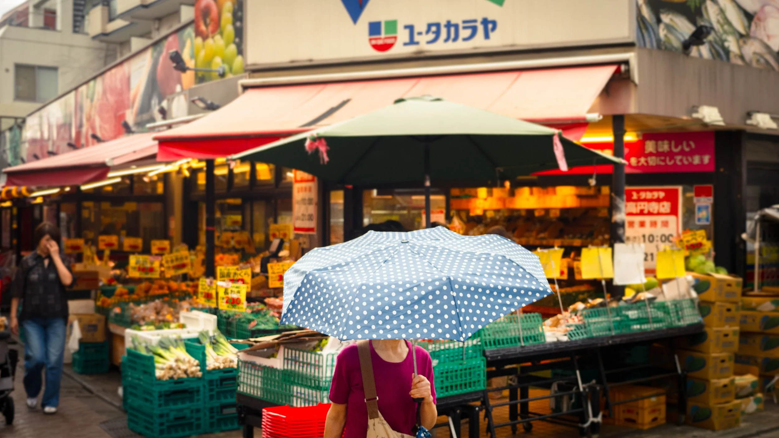 Marché en plein air avec des vendeurs de fruits et légumes sous des parasols variés, une personne en premier plan tient un parapluie à pois blanc et bleu.