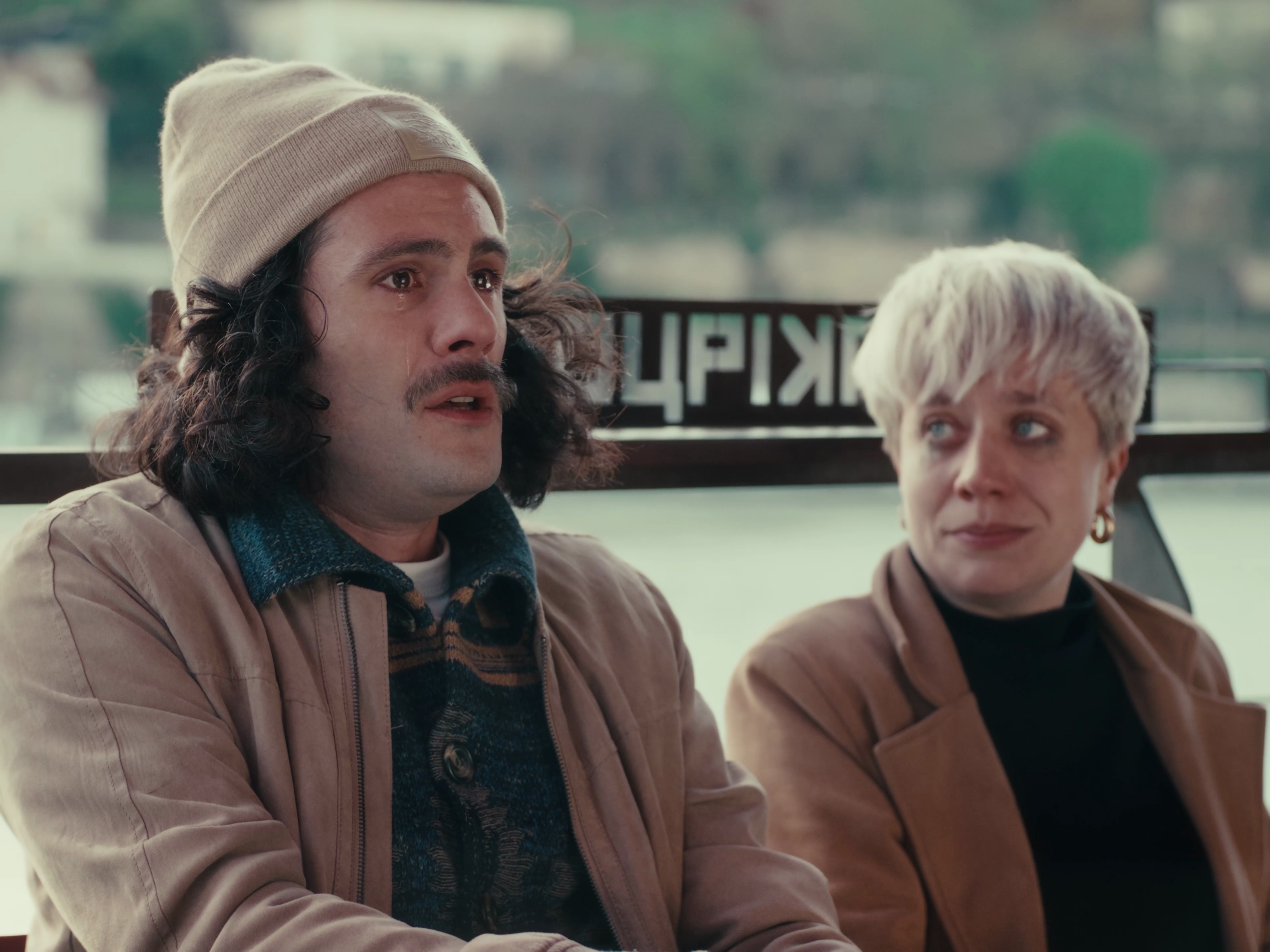 Deux personnes assises à l'intérieur d'un bus ou d'un taxi, une homme avec un chapeau beige et une femme avec des cheveux courts, toutes deux portant des vêtements décontractés.