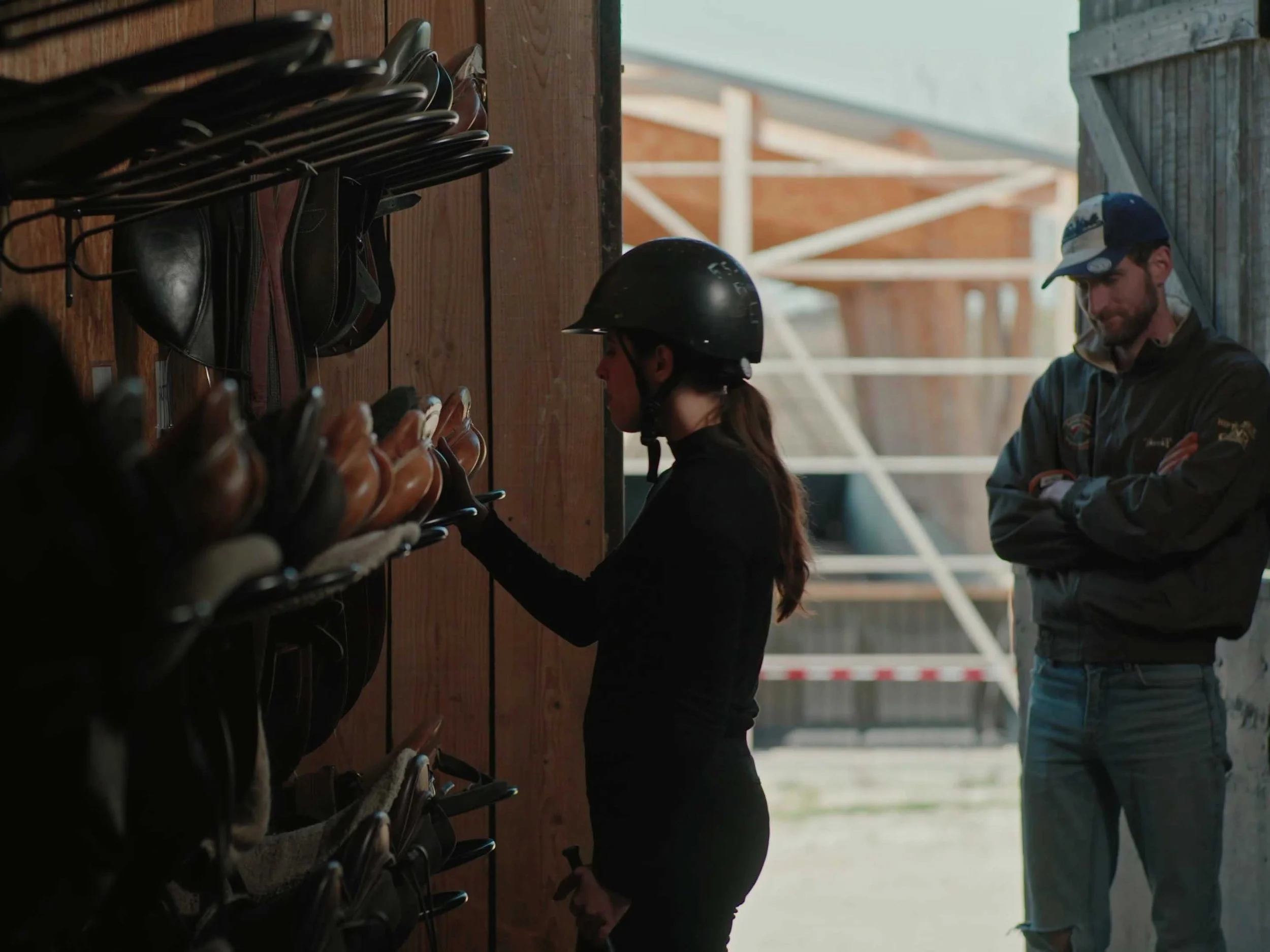 Une femme portant un casque d'équitation regarde des bottes d'équitation sur une étagère en bois, pendant qu'un homme la regarde avec ses bras croisés à côté d'elle