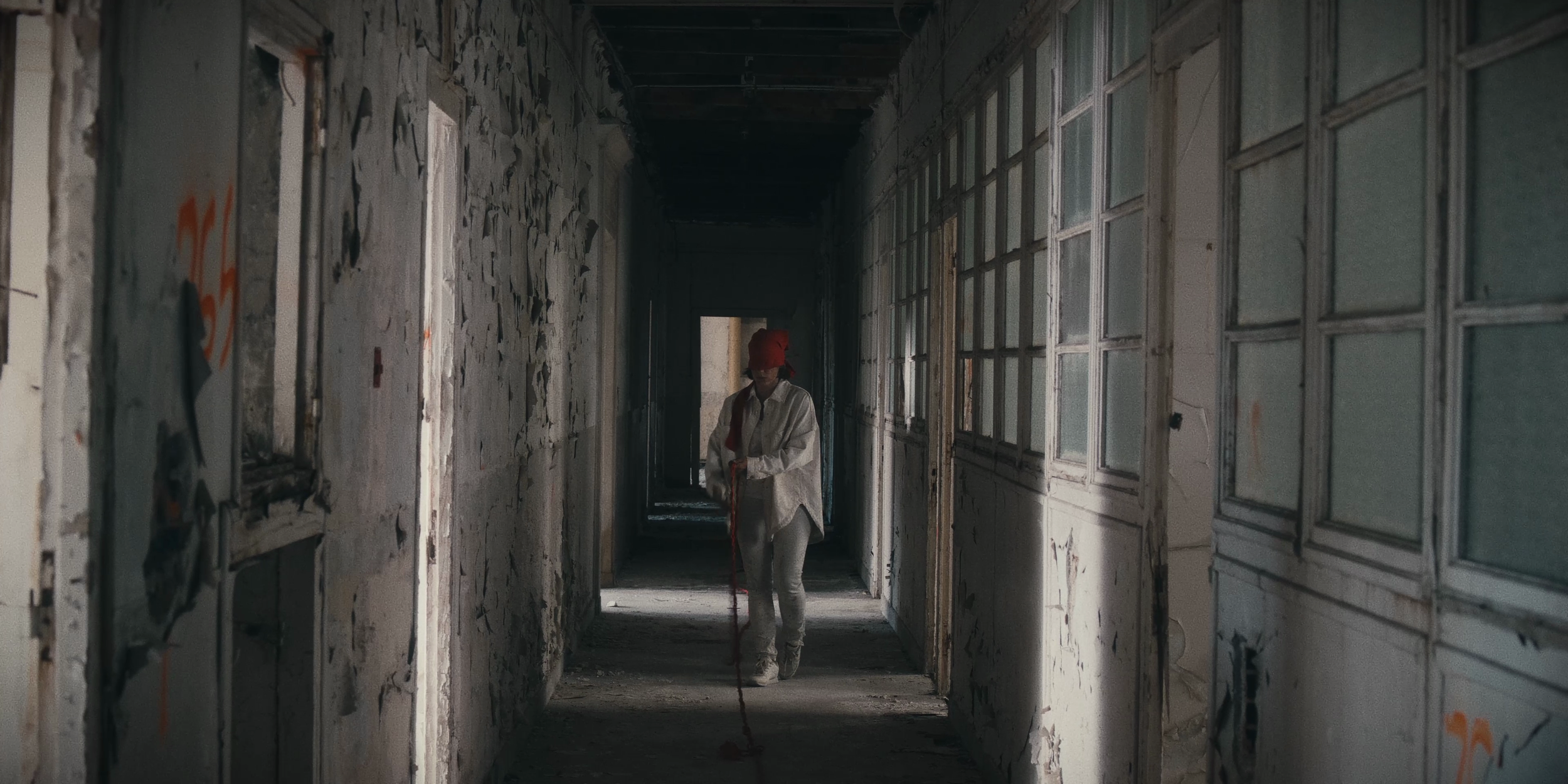 Une femme portant un casque rouge et un vêtement blanc marche dans un couloir abandonné et délabré, avec des murs et des fenêtres endommagés.