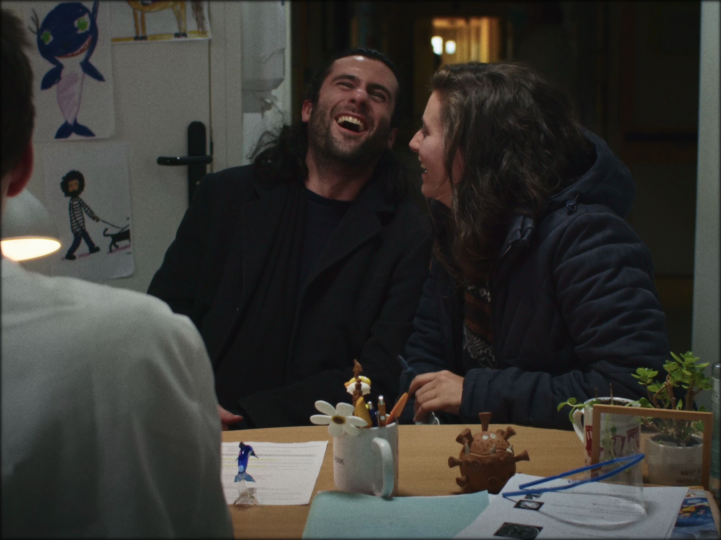 Deux adultes riant ensemble à une table, avec un enfant vu de dos sur la gauche, dans une pièce décorée avec des dessins d'enfants sur le mur.