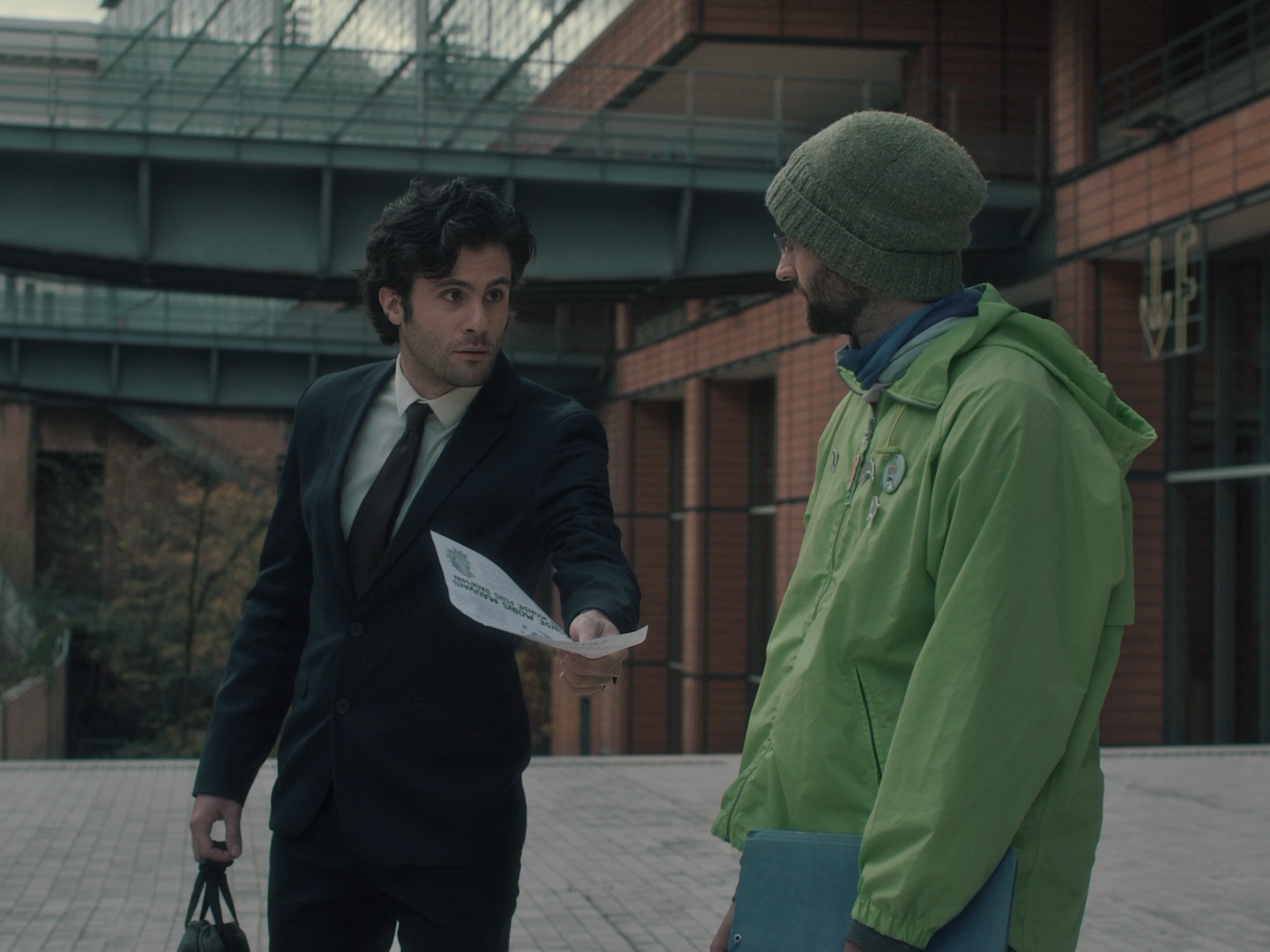 Deux hommes conversent à l'extérieur, un en costume noir avec une ordonnance à la main, l'autre en veste vert olive avec un bonnet, devant un bâtiment moderne.