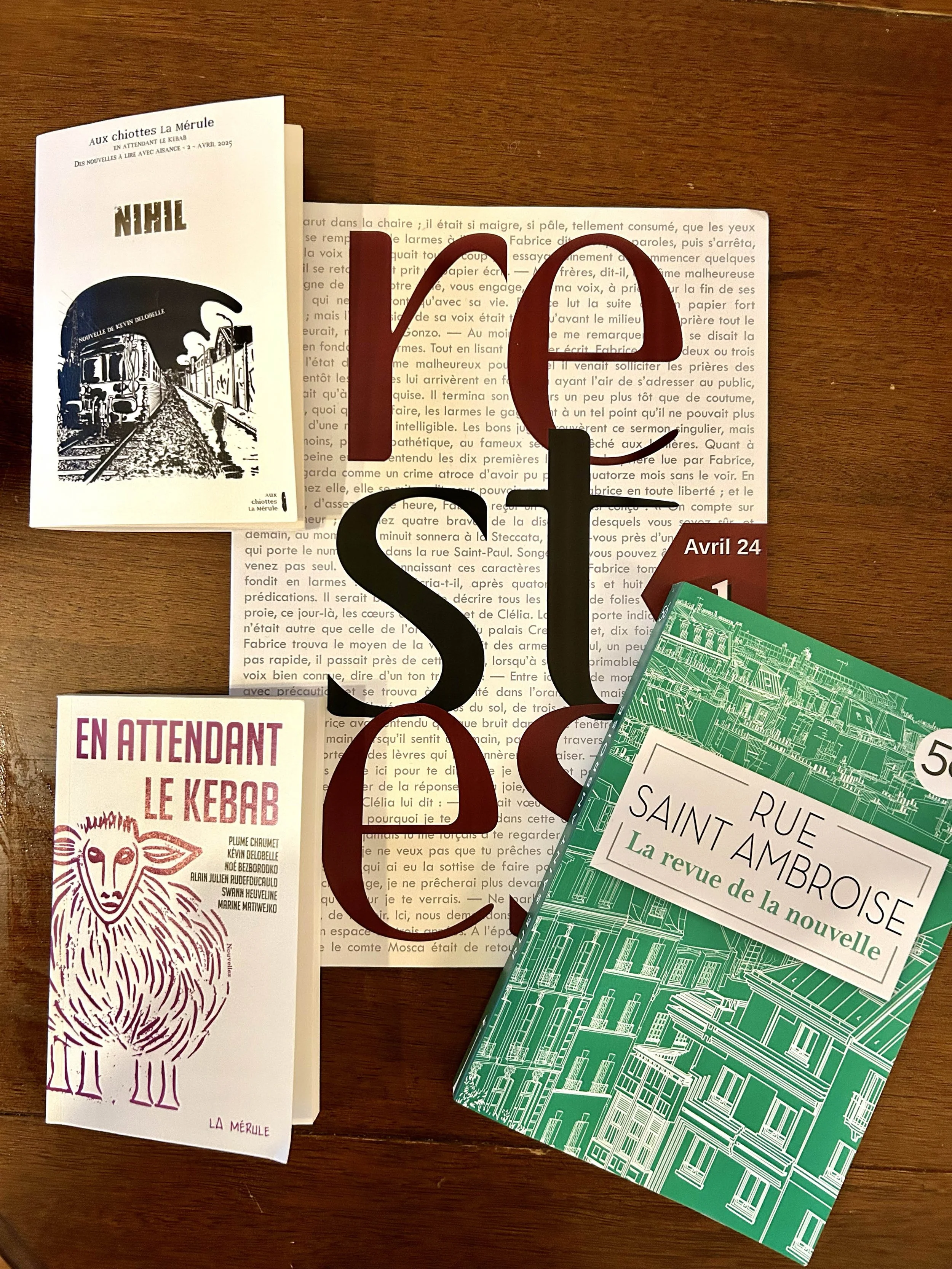 Un livre blanc avec un dessin en noir et blanc d'un train et une personne sur le quai, une brochure violette avec un dessin d'une chèvre et le texte 'En attendant le kebab', un livre vert intitulé 'Rue Saint Ambroise, La revue de la nouvelle', ainsi que des lettres rouge, noir et marron formant le mot 'YES' posé sur une feuille imprimée en français.