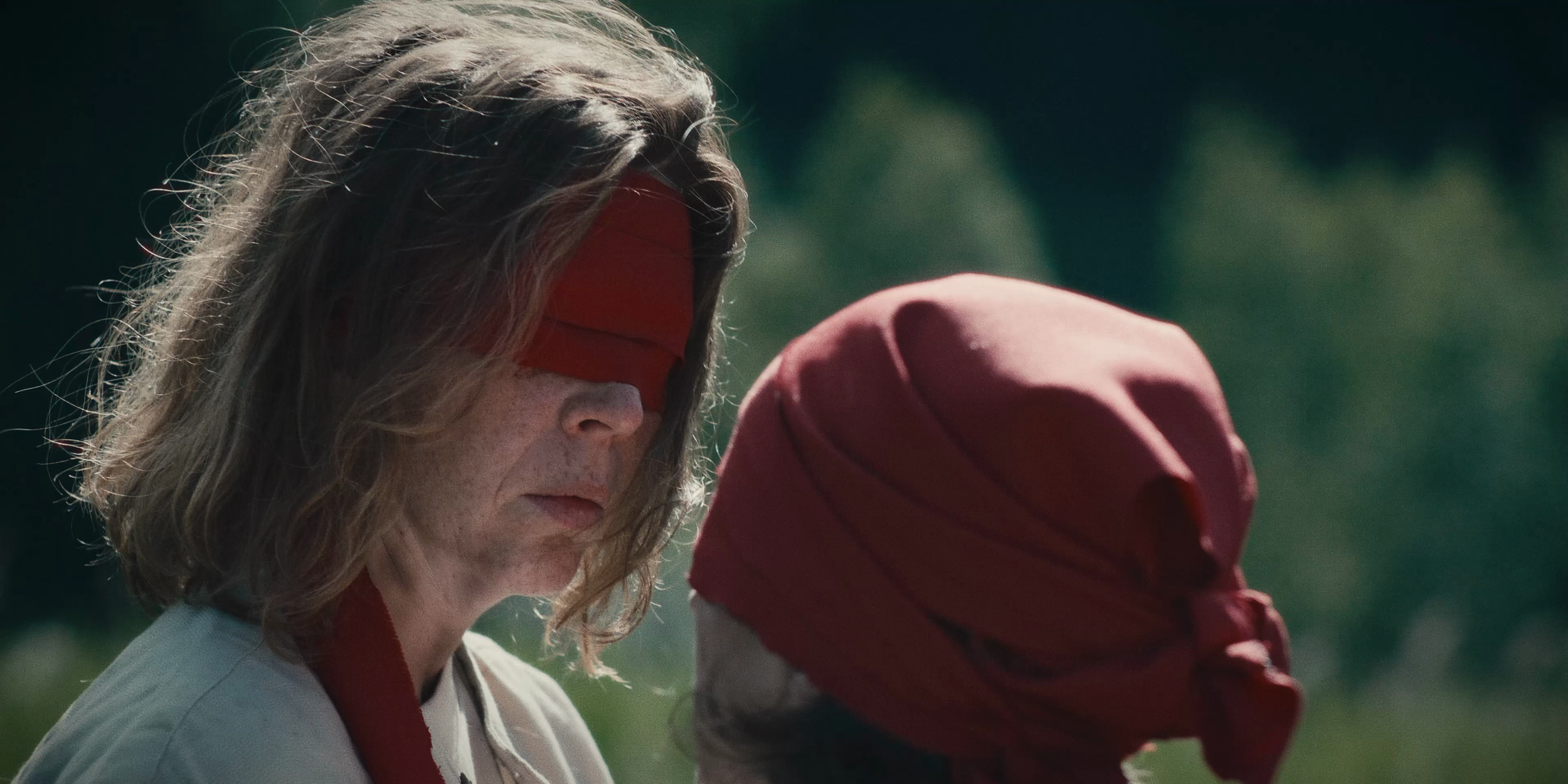 Une femme avec des cheveux longs et bruns, portant un bandeau rouge sur les yeux, se tient près d'une autre personne portant un foulard rouge sur la tête.