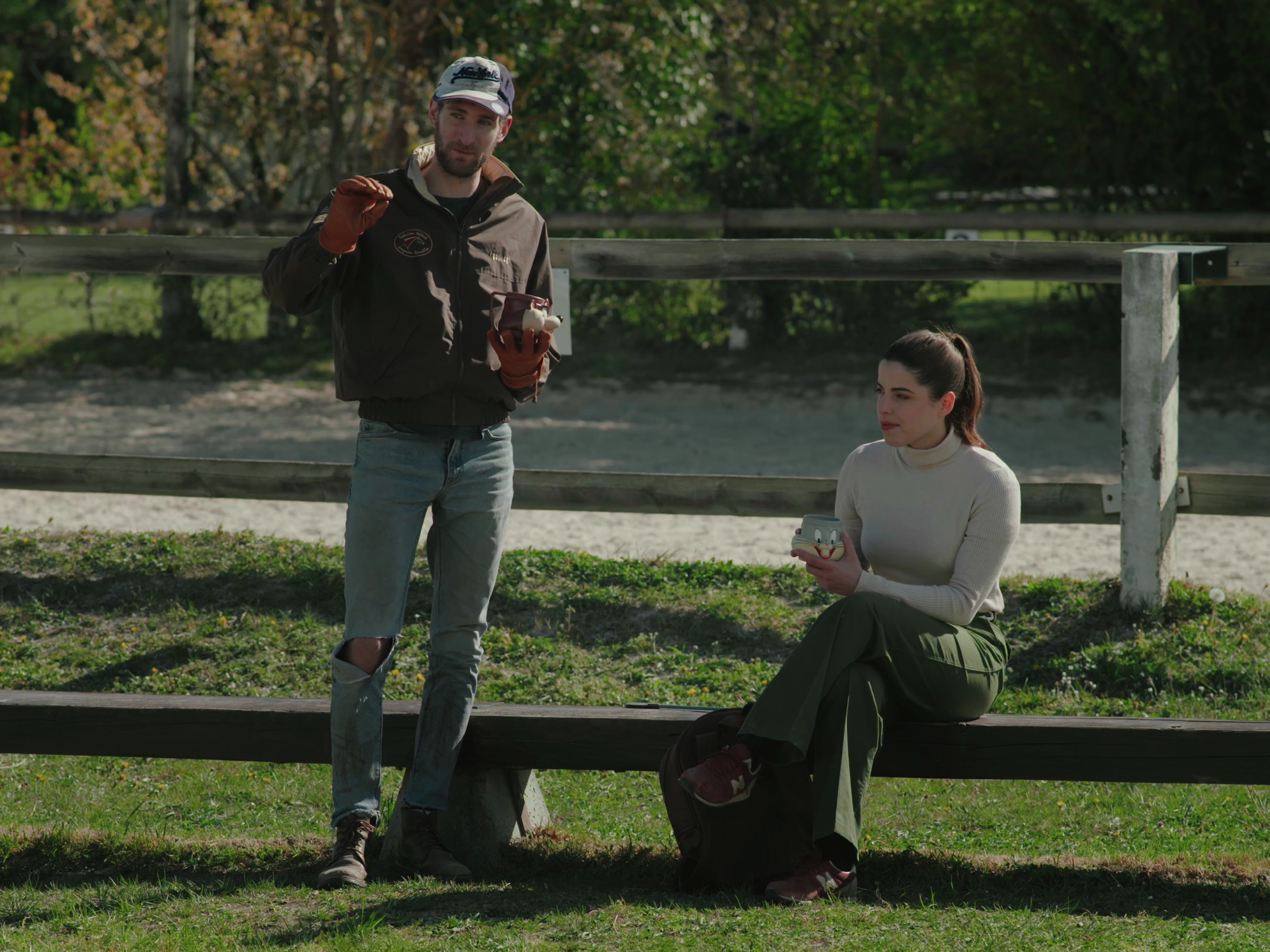 Un homme et une femme dans un parc, avec un mur en bois derrière eux. L'homme, debout, tient un objet dans une main et un autre dans l'autre main, portant une veste brune, un jean déchiré et une casquette. La femme, assise sur un banc, regarde une boîte en forme de visage de cartoon, portant un pull blanc et un pantalon vert.