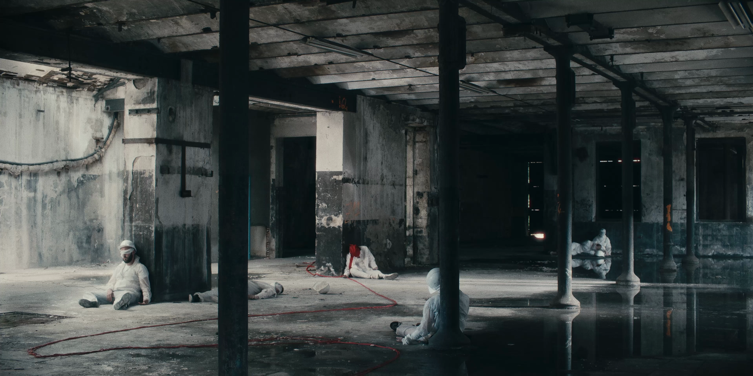 Groupe de personnes en tenue de protection blanche dans un bâtiment abandonné et délabré, assises ou allongées par terre, avec des poutres en métal et des fenêtres cassées.