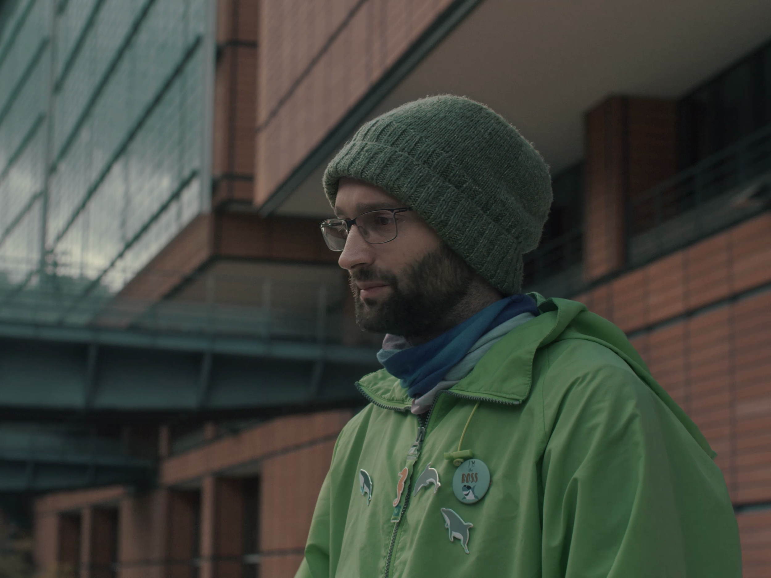 Homme portant un bonnet gris, des lunettes et une veste verte, regardant vers le bas, devant un bâtiment moderne en brique.