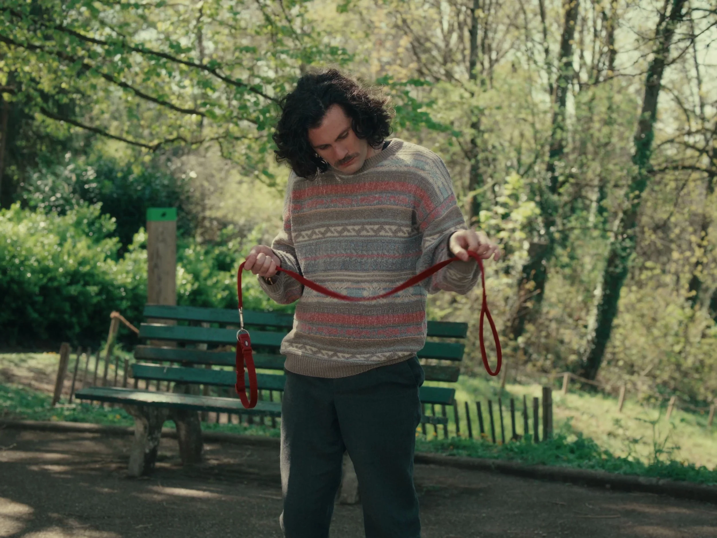 Un homme avec des cheveux bouclés et une moustache, portant un pull à motifs, tient une laisse rouge dans un parc verdoyant.