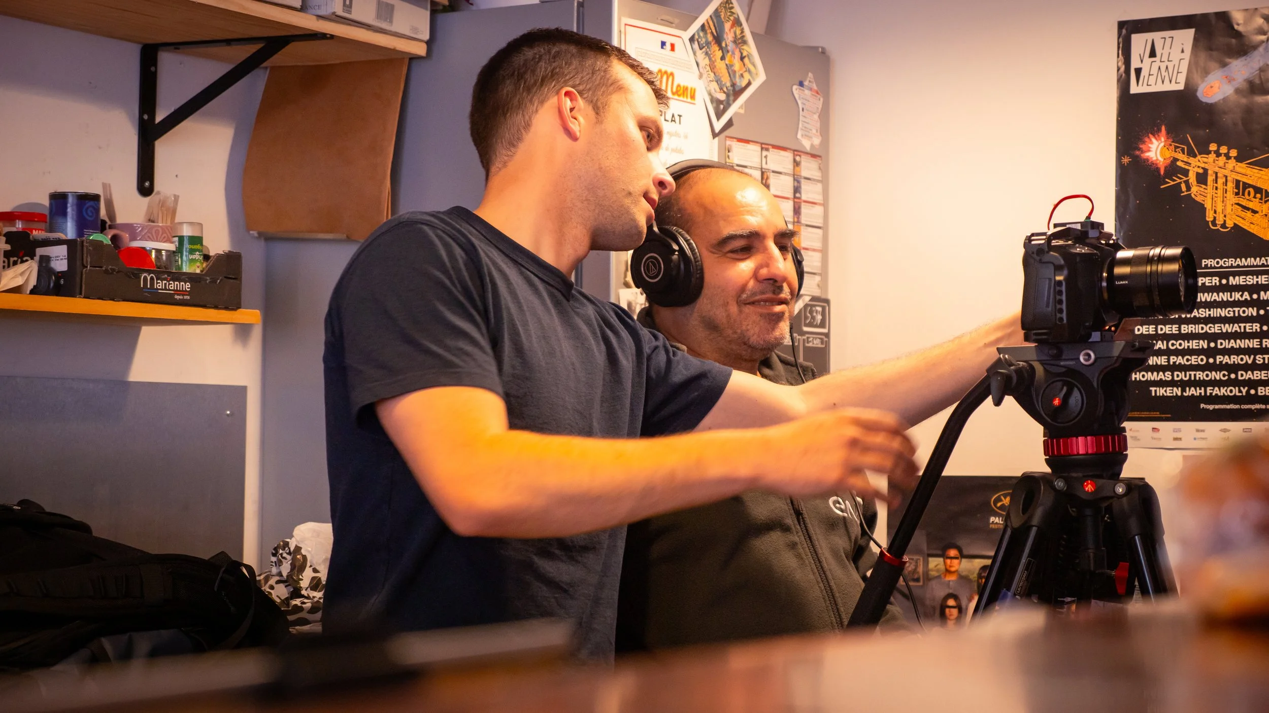 Deux hommes ajustent une caméra sur un trépied dans une pièce. Un homme porte un casque audio.