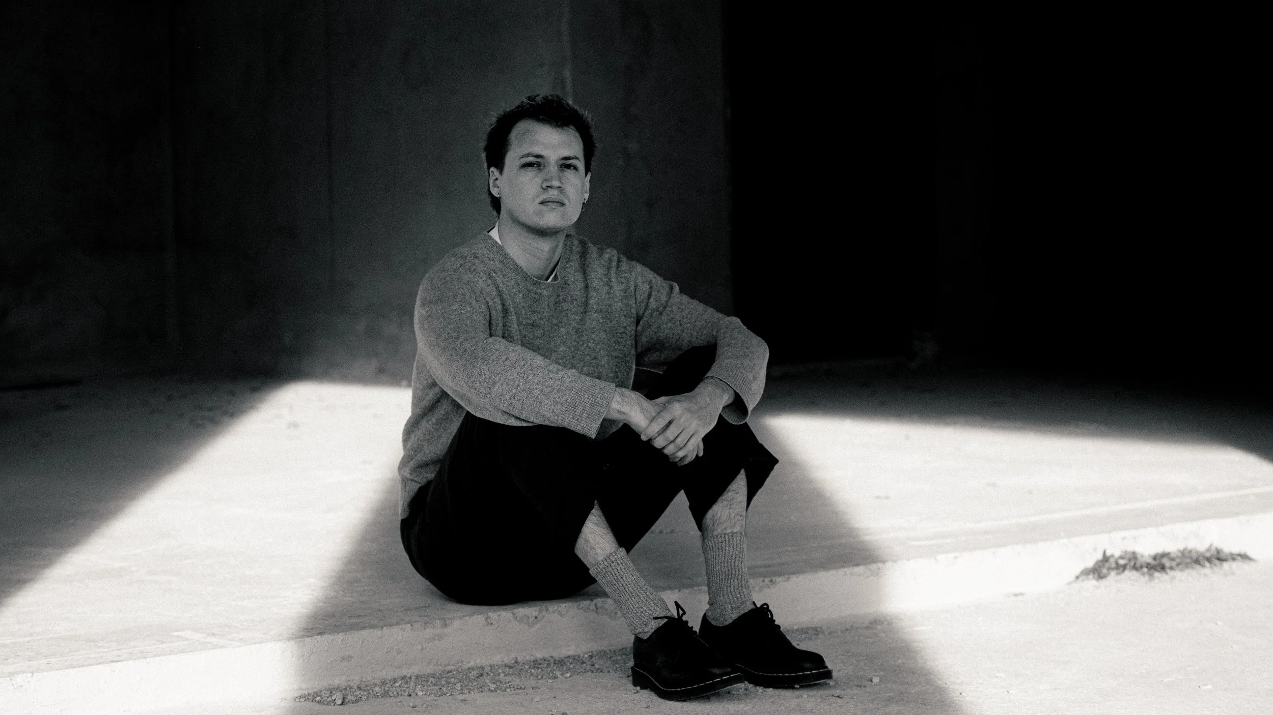 Un jeune homme assis au sol dans une pièce sombre, avec un éclairage créant un contraste marqué. Il porte un pull gris, un pantalon noir, des chaussettes épaisses et des chaussures noires.