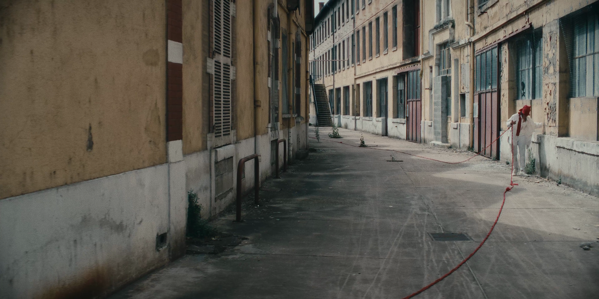 Une personne en blanc et rouge, portant un chapeau rouge, marche dans une ruelle bordée de bâtiments anciens avec des fenêtres et des portes rustiques, tenant une corde
