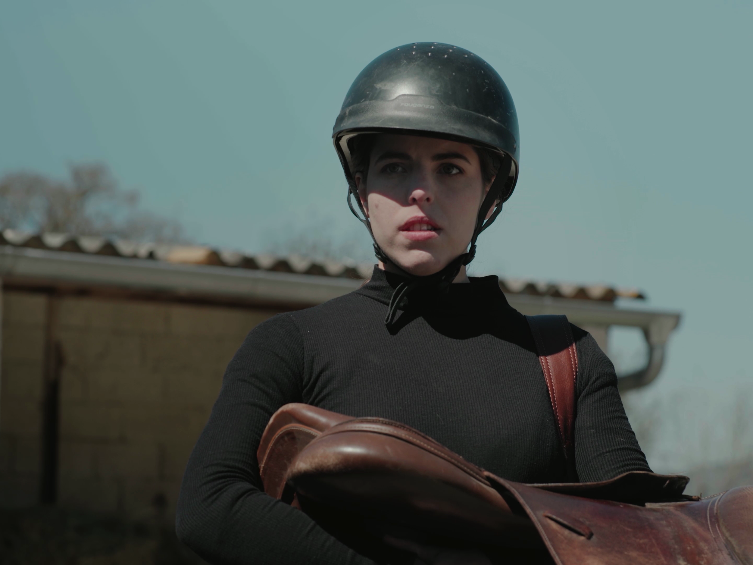 Jeune femme portant un casque de moto, tenant un sac en cuir, à l'extérieur, avec un bâtiment derrière elle.