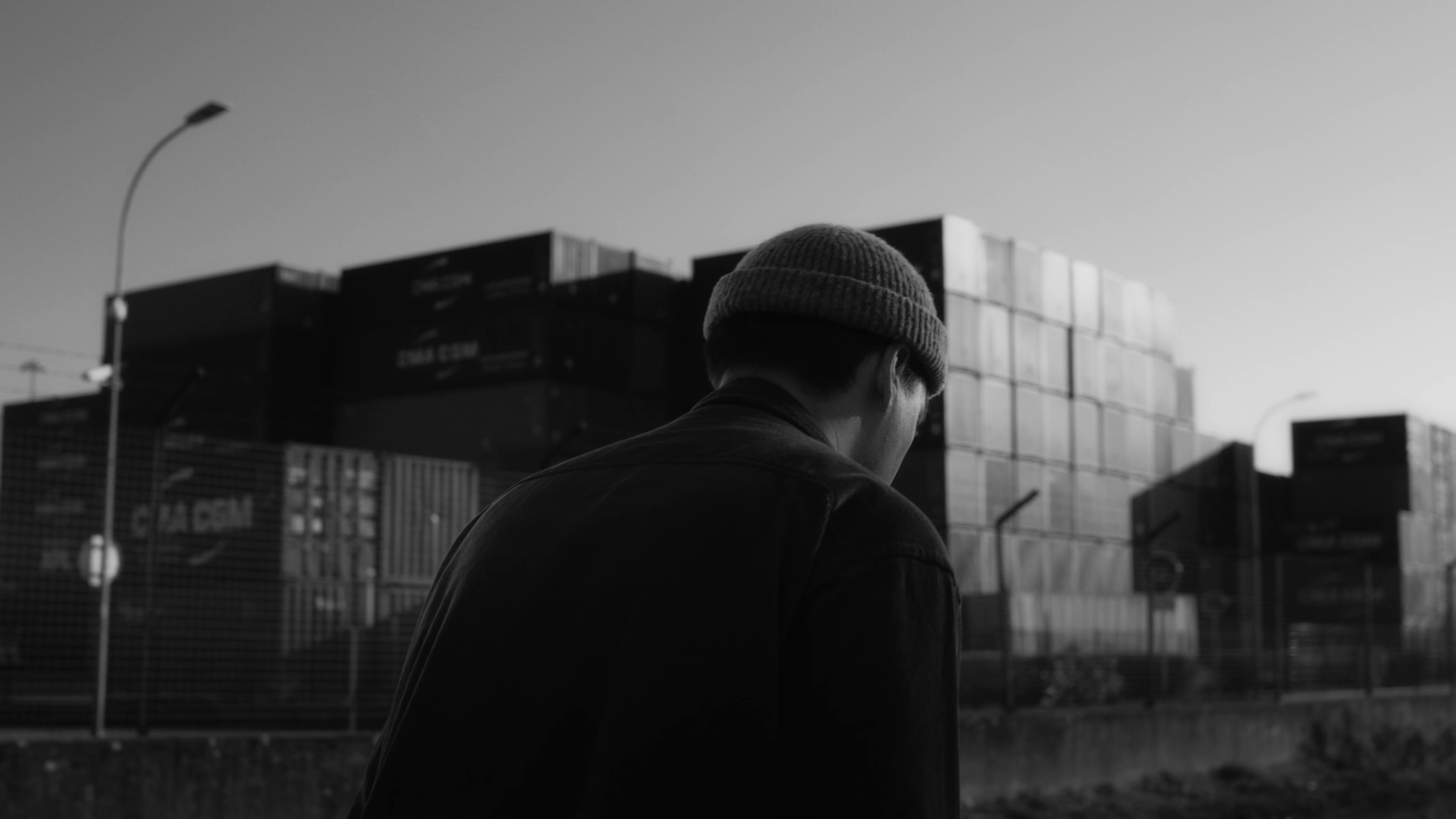 Un homme portant un bonnet regarde vers l'avant dans un environnement industriel avec des containers en arrière-plan.