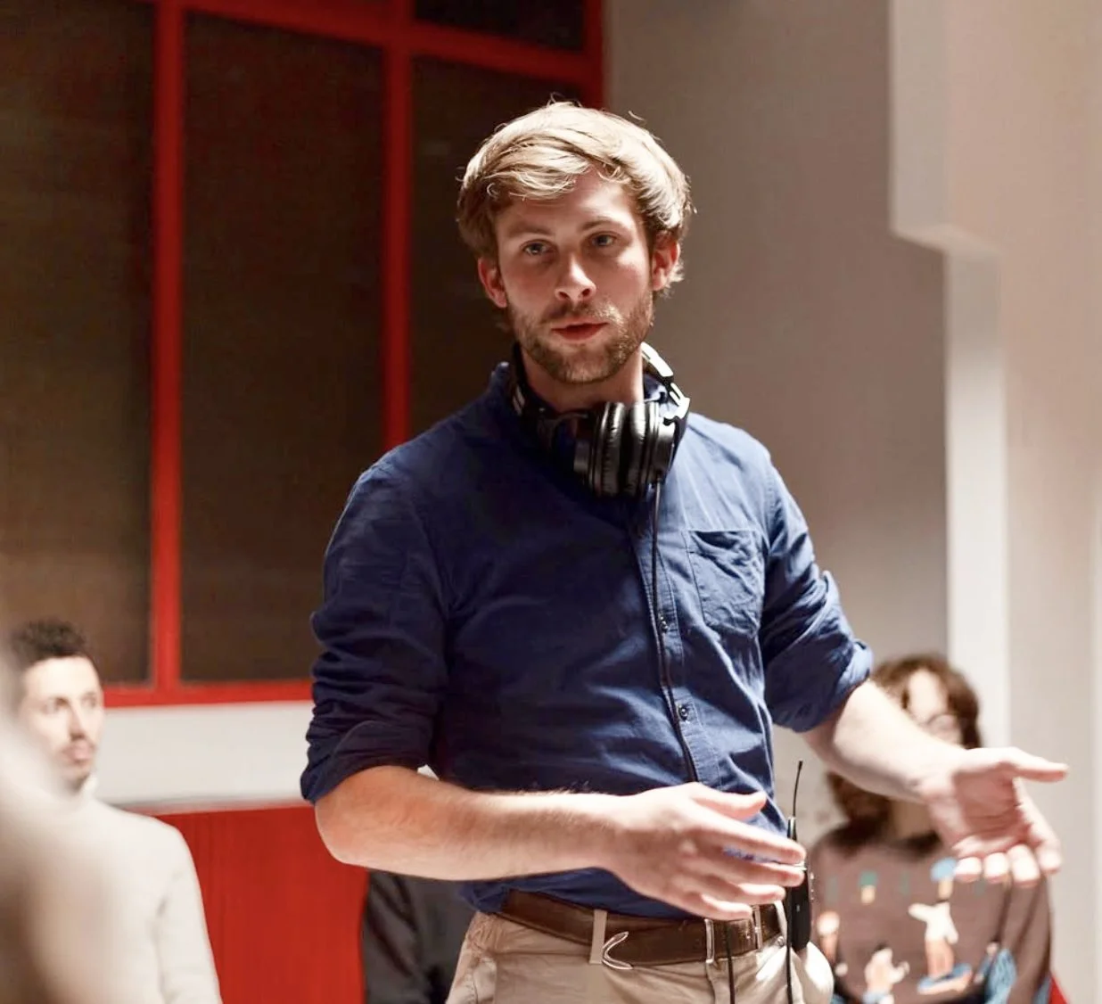 Un homme avec des cheveux châtains, une barbe légère, portant une chemise bleue à manches longues et des écouteurs autour du cou, parle lors d'une réunion ou d'un atelier.